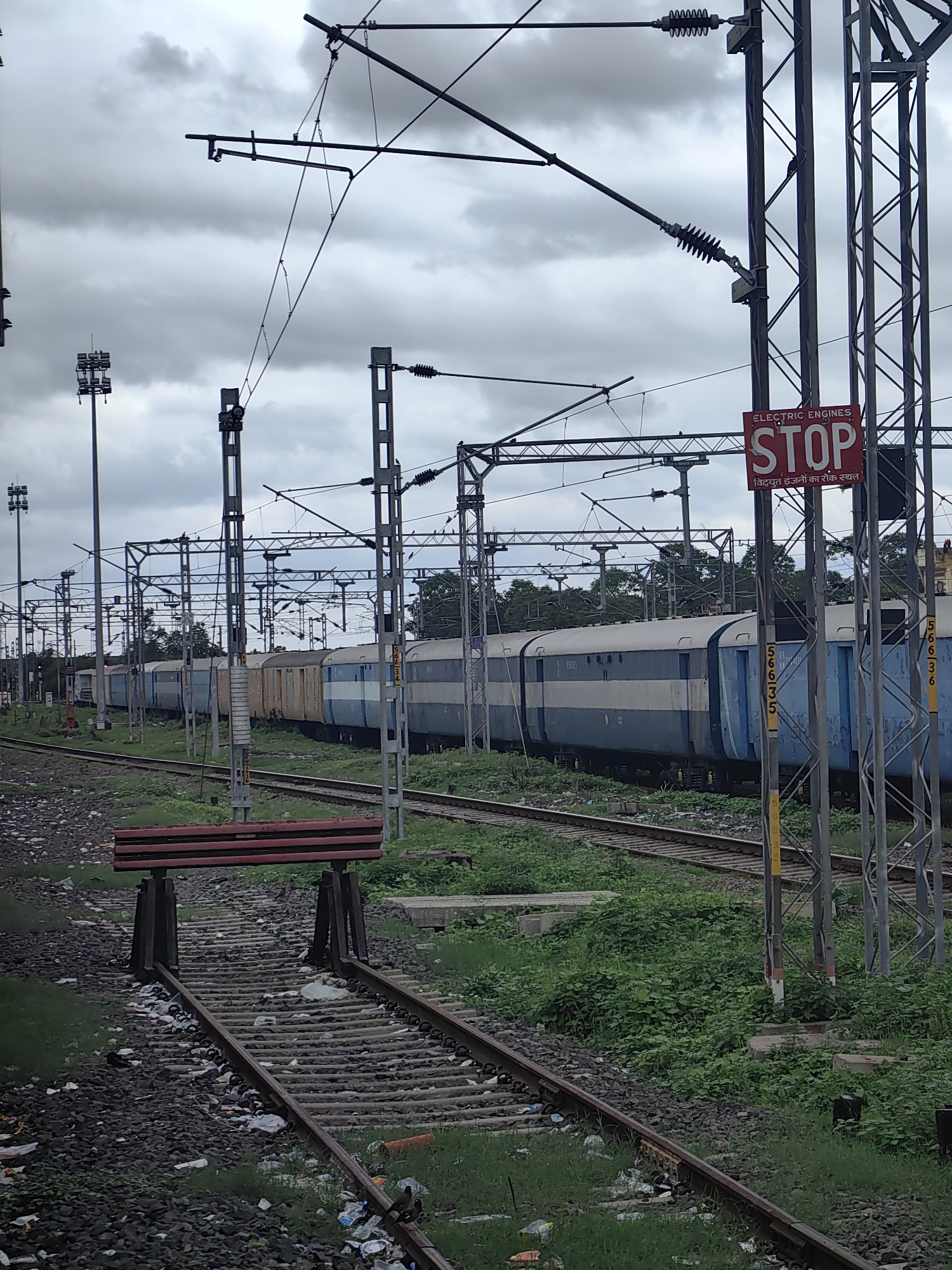 An overcast day frames a railway yard in Khandwa, India, where a long train with blue and white carriages is partially visible on the tracks. Overhead, a complex network of power lines for electric trains crisscrosses the sky, supported by metal poles. A red and white "STOP" sign, with text in English and Hindi, is prominently displayed on one of the poles, indicating a restriction for electric engines. The foreground shows a set of railway tracks littered with debris, with a simple red and black buffer stop at the end of one set of tracks. Lush green vegetation grows alongside the tracks, contrasting with the industrial elements of the scene. The overall impression is one of an active, yet slightly neglected, railway infrastructure.