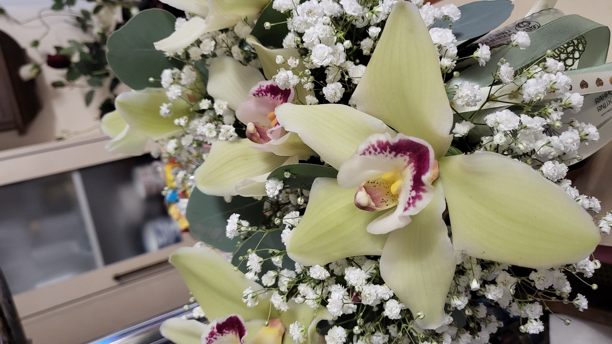 A close-up shot of a bouquet of light green orchids with deep purple markings. The orchids are interspersed with delicate white baby's breath flowers and some muted green eucalyptus leaves. The bouquet is tied with decorative ribbons, one of which features a repeating pattern of small gold hearts and text. The background is softly blurred, showing a hint of cabinetry and possibly a shelf with items. The image does not contain any elements that suggest a location within the city of Chaikovsky, Russia.