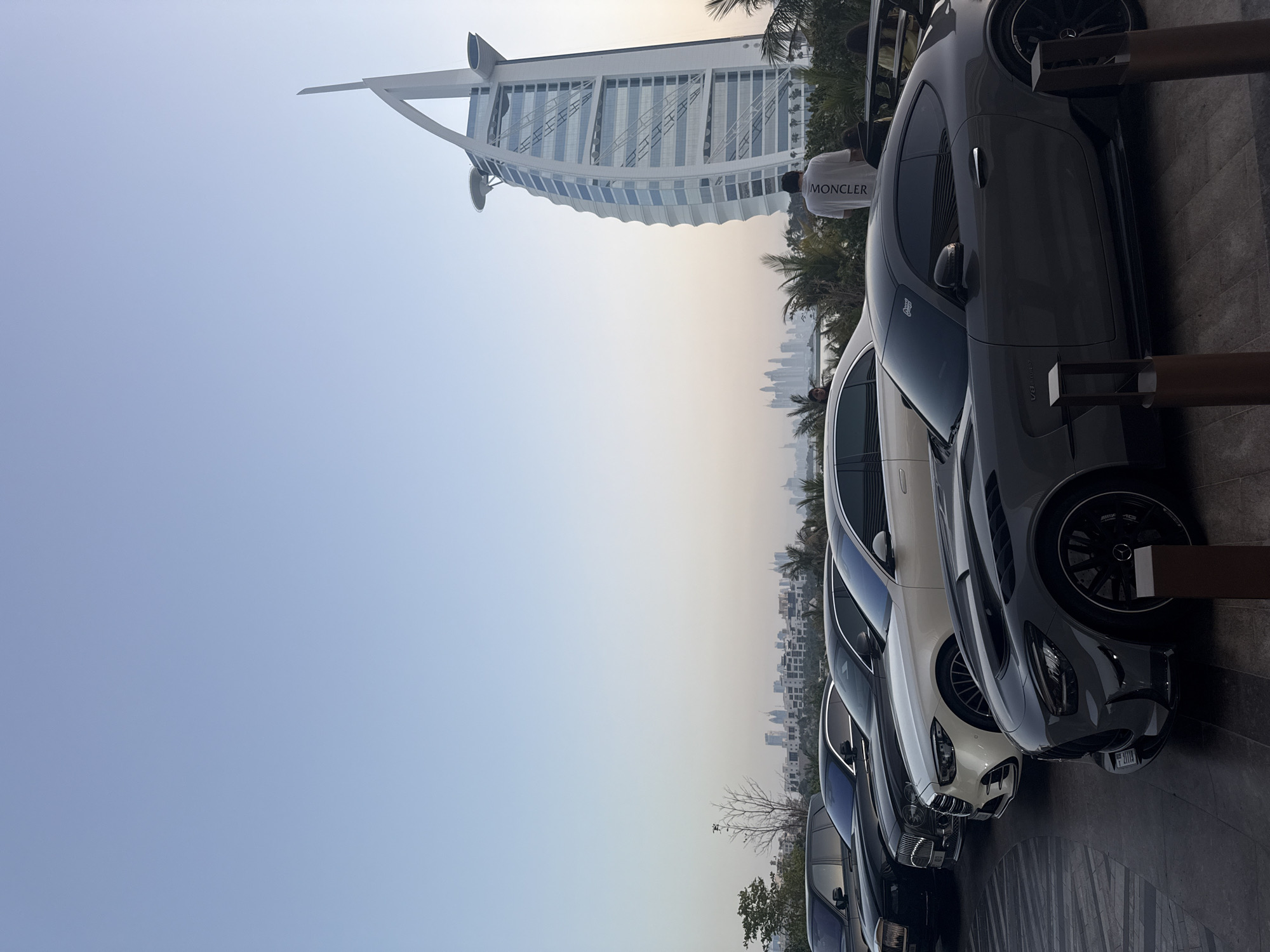 The image captures a row of luxury cars parked in front of the Burj Al Arab hotel in Dubai, United Arab Emirates. The distinctive sail-shaped architecture of the hotel is visible in the background. Beyond the hotel, the Dubai skyline appears hazy in the distance. A person wearing a "MONCLER" shirt is standing near the cars. The cars are parked on a paved area, likely a parking lot. The sky is a gradient of light blue to a hazy sunset color.