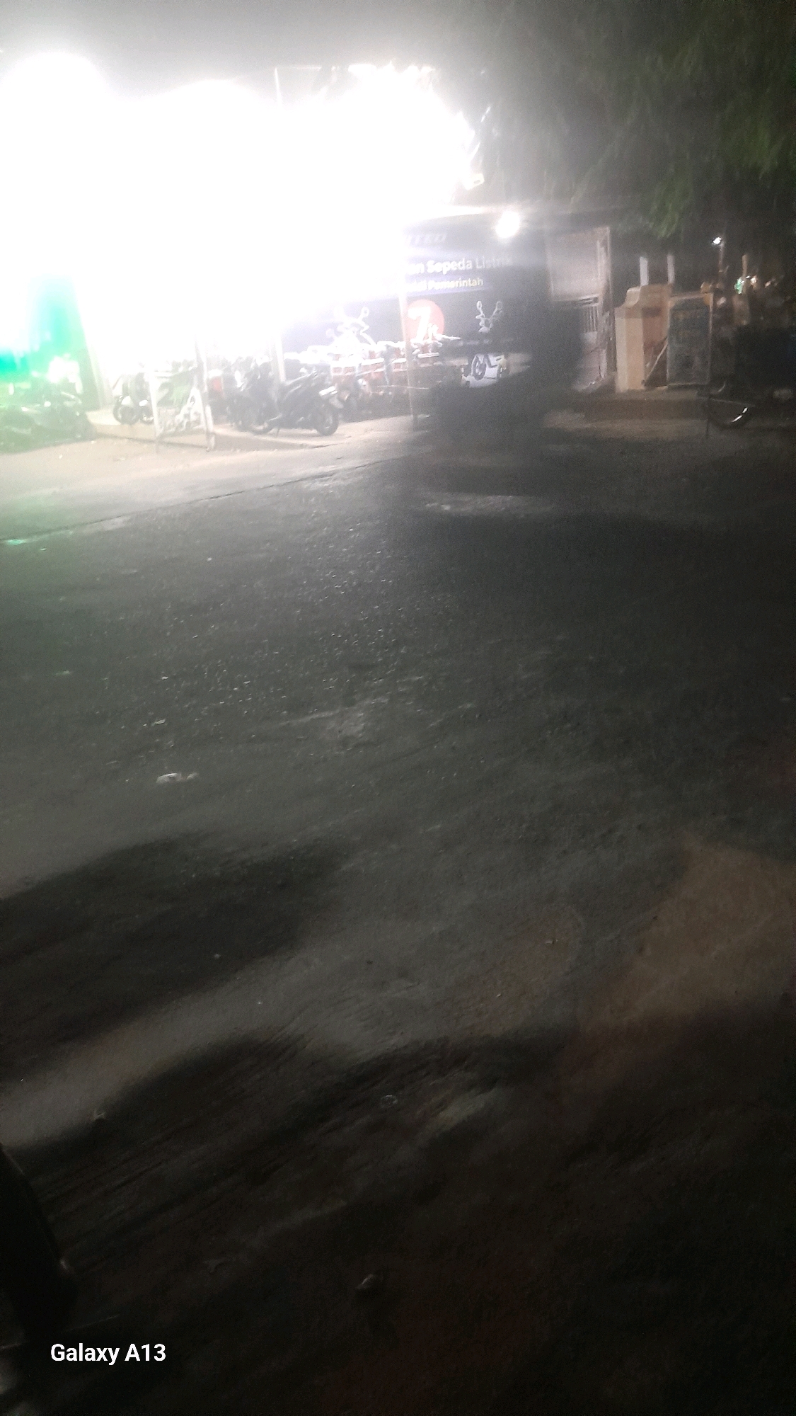 This image depicts a nighttime scene on a paved road in Jekulo, Indonesia, likely near a business or establishment. A bright, overexposed light source dominates the upper left portion of the frame, obscuring details in that area. To the right of the bright light, a storefront or building is visible, with illuminated signage. Text on the sign, though partially obscured and blurry, appears to read "Sukan Sepeda Listrik" and "Pemerintah". A number of motorcycles are parked in front of the establishment, and some appear to be electric scooters. A person is partially visible in the lower left corner of the image. The road surface is dark and appears wet or reflective, with visible tire marks and scattered debris. Shadows are cast across the road, suggesting the presence of unseen objects or structures. The overall impression is that of a somewhat gritty urban environment at night.