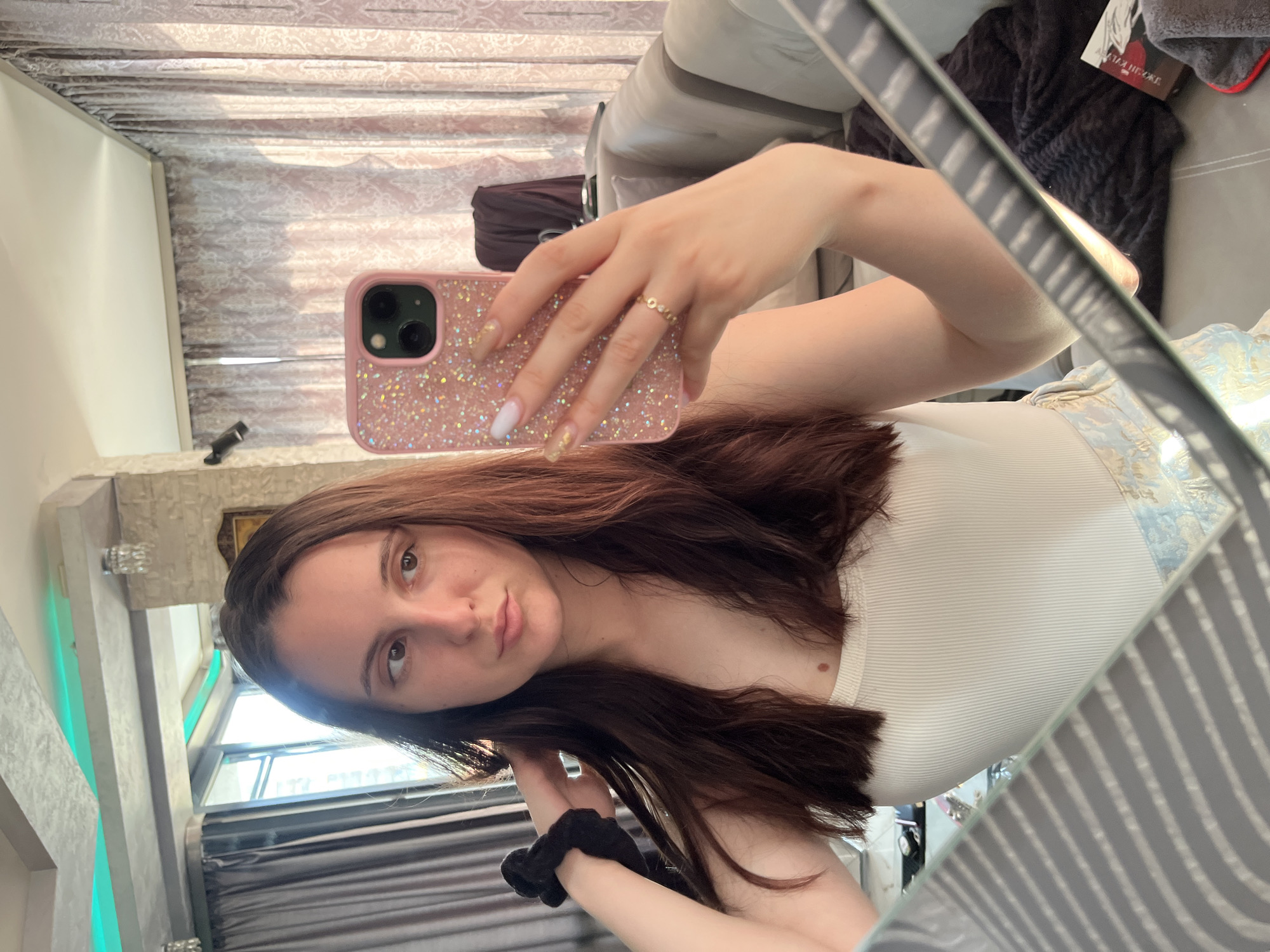 A sideways mirror selfie captures a young woman in an indoor setting, likely in Pleven, Bulgaria. The woman, with long dark brown hair, fair skin, and brown eyes, wears a white ribbed tank top and looks directly at the camera with a calm, direct expression.
She holds a pink smartphone with a glittery case in her right hand, with a delicate gold-colored ring on her right ring finger. Her nails are manicured with a mix of nude/beige, white, and gold-flecked polish. A black scrunchie is on her left wrist.
The reflected background reveals a domestic interior, possibly a living room or bedroom. Elements include patterned textile blinds, a dark grey textured blanket, and a light-colored sofa. A decorative wall feature with stone-like texture and green ambient lighting is visible. Natural light coming through a window indicates it is daytime. A partial word, "AKAIL...", is faintly discernible on a book or magazine.