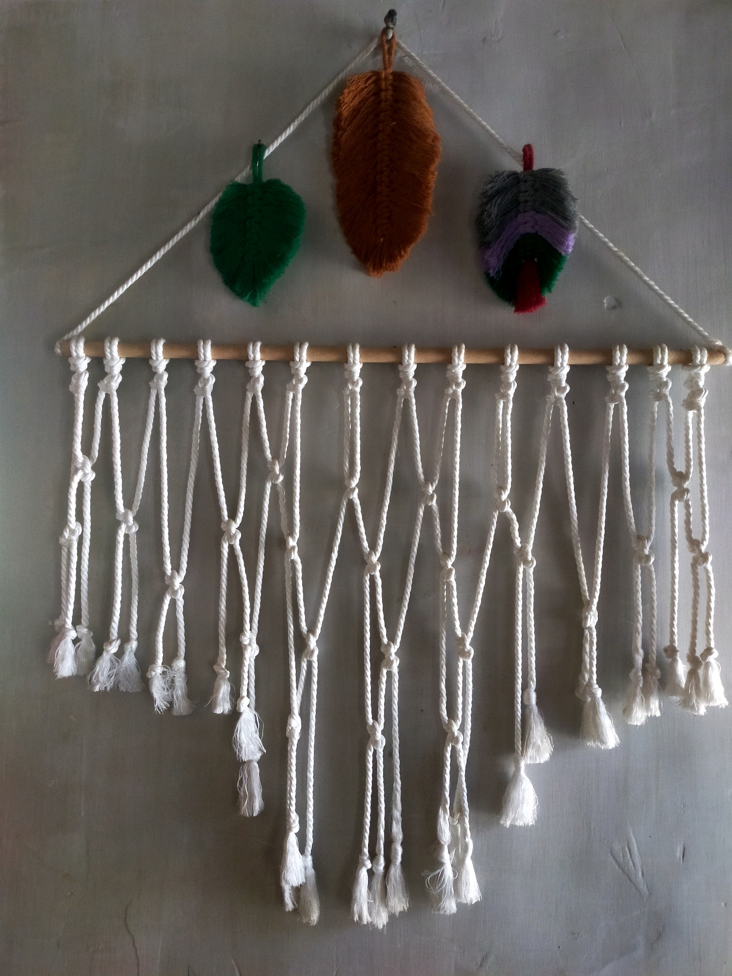 A macrame wall hanging is displayed against a textured gray wall. The wall hanging is suspended from a wooden dowel rod and features a triangular arrangement of three decorative leaf-shaped ornaments at the top. The leftmost leaf is green, the middle leaf is brown, and the rightmost leaf is a multi-colored feather-like ornament with gray, purple, green, and red segments. Below the dowel rod, numerous white macrame cords hang, forming a patterned fringe with various knotting techniques and ending in tassels at different lengths. The setting is indoors, with the gray wall suggesting a domestic or workshop environment. The image is a static, top-down shot of the wall hanging.