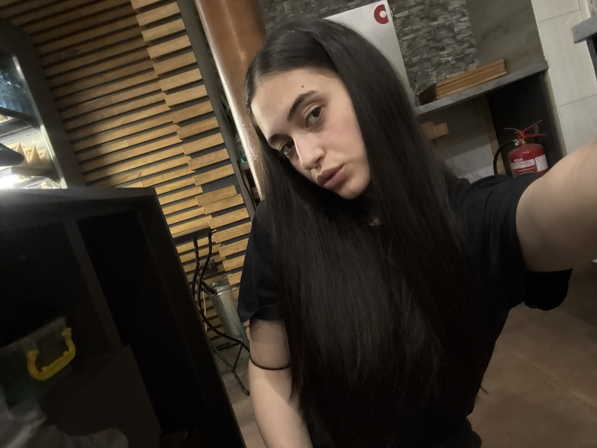 The image is a selfie of a young woman with long, dark hair, wearing a black t-shirt. She is indoors, in what appears to be a commercial or industrial space. The background shows a wall with horizontal wooden slats, a metal pillar, and some shelves with items on them, including a red fire extinguisher. The floor is made of brown tiles. The lighting is dim, with a light source coming from the left. The woman is looking directly at the camera with a neutral expression. The overall impression is of an informal snapshot taken in a utilitarian setting. The location is not specified in the image itself, and thus cannot be determined.