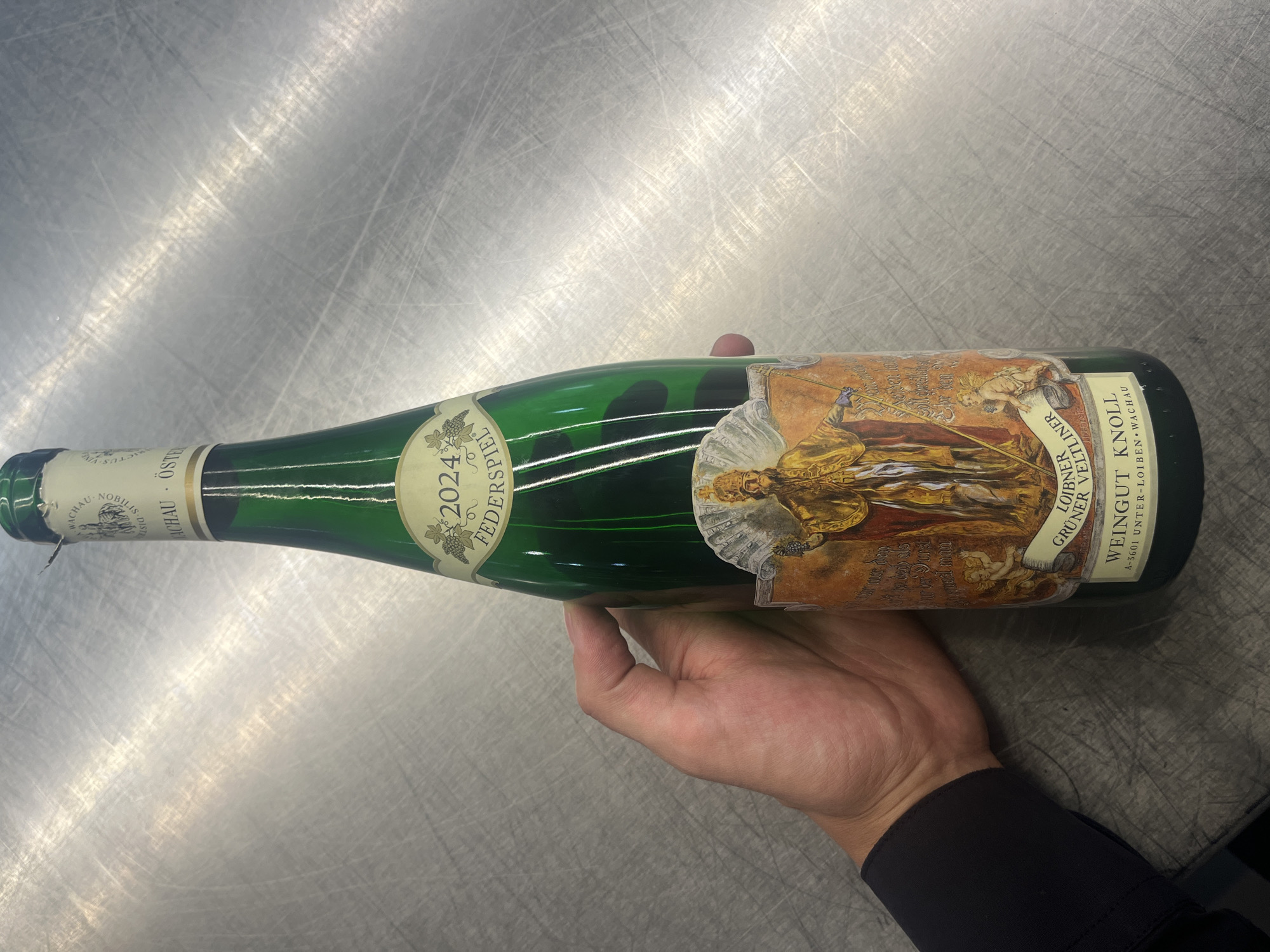 A close-up, eye-level shot captures a hand holding a green wine bottle horizontally over a brushed metal surface. The bottle is the primary focus, cradled by a person's right hand, which wears a dark long-sleeved garment, possibly a shirt cuff.
The bottle is made of dark green glass and features two distinct labels. The main label is ornate and richly detailed, predominantly in hues of gold, brown, and cream. It depicts a regal figure, possibly a saint or monarch, holding a staff, surrounded by intricate designs and cherubic figures. Text visible on this main label includes "LOIBNER", "GRÜNER VELTLINER", "WEINGUT KNOLL", and "A-3601 UNTER-LOIBEN WACHAU", indicating the wine's origin is Austria's Wachau region.
A smaller, lighter-colored neck label is also visible, prominently displaying "2024", likely the vintage year. Other legible text on this label includes "FEDERSPIEL", "WACHAU", and parts of "ÖSTER", further confirming the Austrian origin. The foil or capsule covering the cork is a light, possibly white or cream, color.
The background is a textured, reflective metal surface, likely a countertop or table, showing numerous fine scratch marks and reflections from overhead lighting. The activity is simply presenting or holding the bottle. No specific time of day or weather is discernible. The labels provide strong geographical cues, pointing to Austria, which contradicts the provided general location context of Sofia, Bulgaria. No emotions are expressed by the partially visible hand.