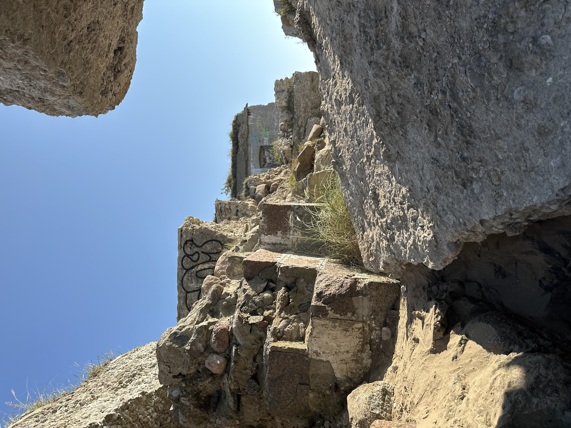 The image depicts the ruins of a coastal fortification under a clear blue sky. The structures are made of rough stone and concrete, showing signs of decay and neglect. Graffiti is visible on some of the walls, suggesting recent human activity. Overgrown vegetation, such as grass and weeds, sprouts from cracks and edges of the ruins. The foreground is dominated by textured sand and rocks, with a deep crevice cutting through the terrain. In the background, a section of a building with a doorway and windows can be seen, also in a state of disrepair. The scene is part of the Liepaja Northern Forts in Liepaja, Latvia, a historical military complex located along the Baltic Sea coast.