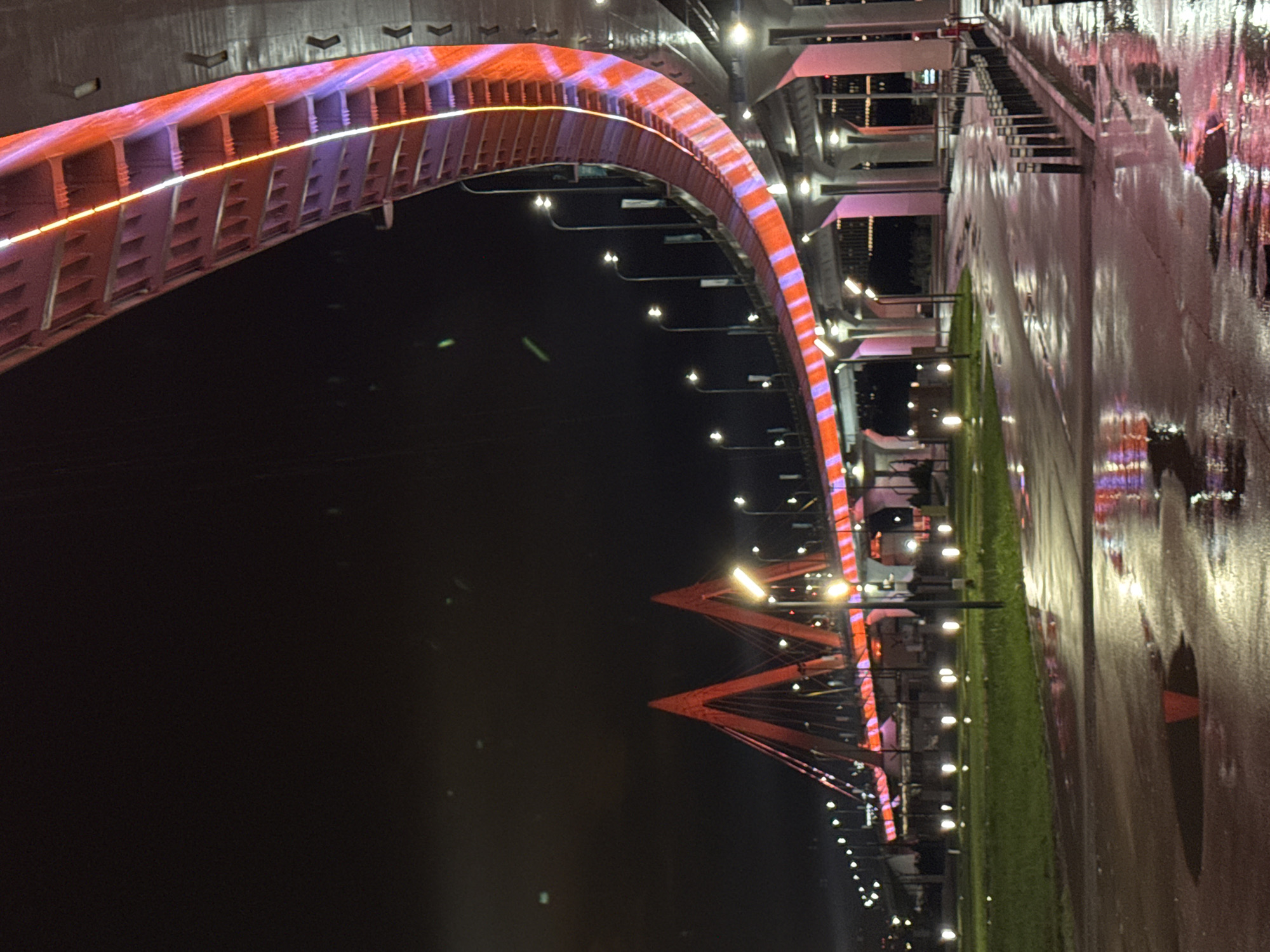 Captured at night, the image showcases the Millennium Bridge in Kazan, Russia. The bridge's arch spans across the dark water, illuminated with a pattern of orange and purple lights. The supports of the bridge can be seen in the distance. On the right side of the image, the landscape transitions from a strip of green grass to a wet, reflective paved area. Streetlights are evenly spaced along the pavement, casting reflections on the wet surface. Buildings are visible in the background. The scene is devoid of people.