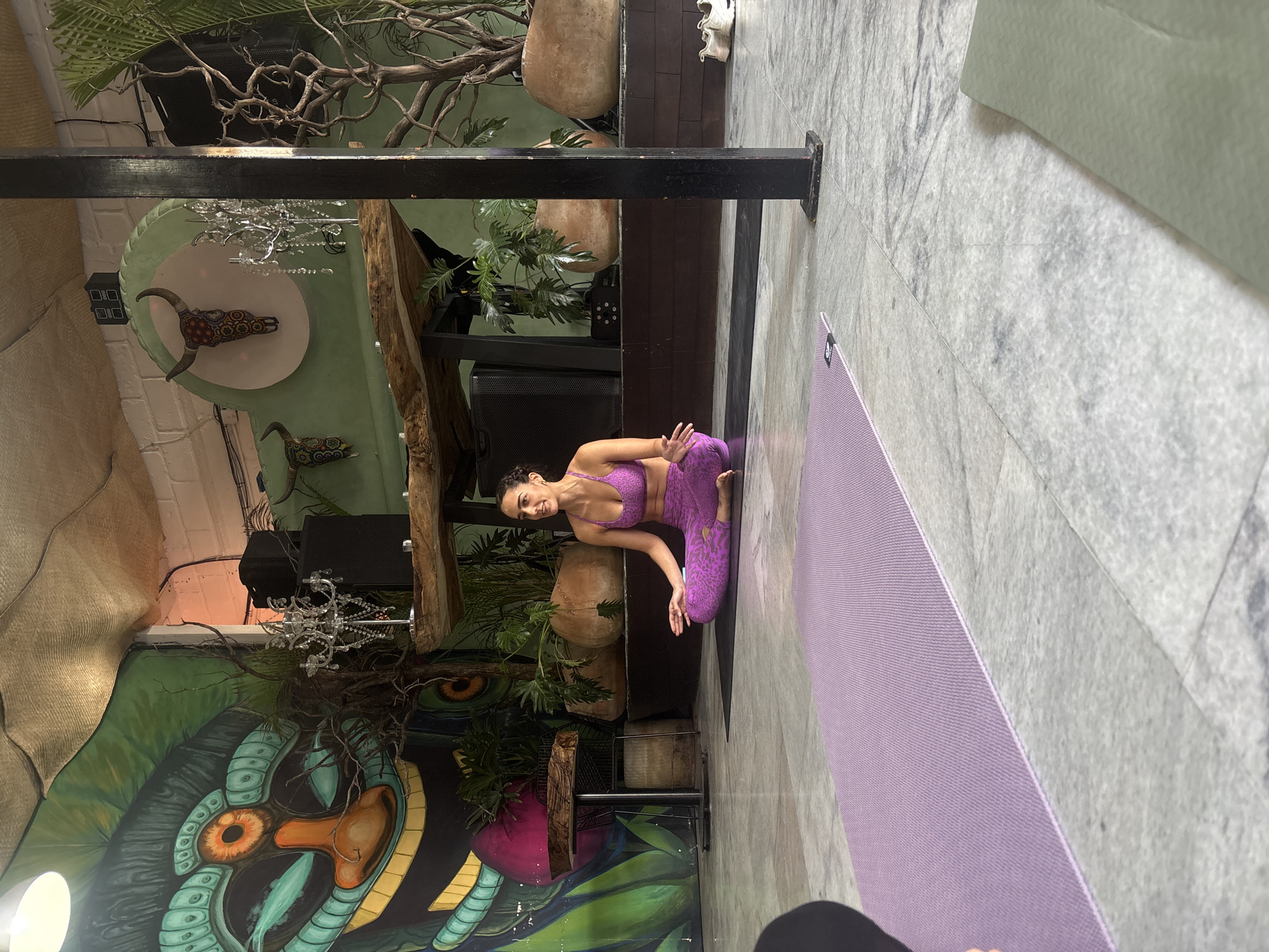 A woman in a purple leopard-print workout set kneels in a yoga pose on a purple yoga mat in a room with eclectic decor. The room features a large, colorful mural of a snake-like creature on the wall, various plants, and a faux chandelier. The location is likely in Cuernavaca, Mexico, given the presence of a mural.