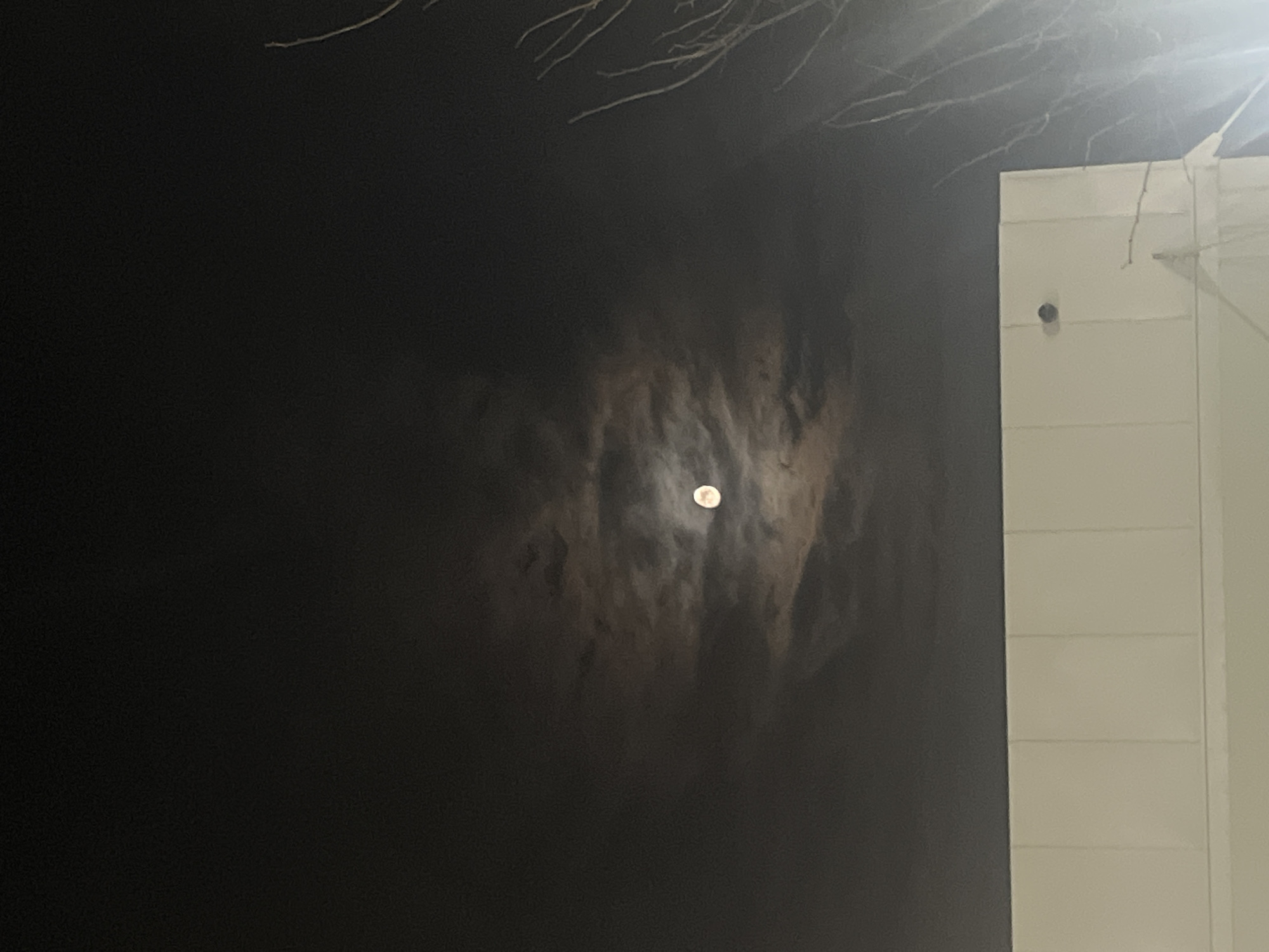 A low-angle, nighttime view depicts a partially obscured moon with a white exterior wall of a building on the right. The moon is a small, round, bright spot surrounded by a halo of diffused light, likely due to atmospheric conditions. Dense, dark clouds or haze partially conceal the moon. Above the moon, thin branches are visible against the dark sky. The wall on the right is pale, with horizontal lines, and a dark, round object (possibly a security camera or light fixture) is mounted on it. The environment is dark and murky, indicative of nighttime. No people or other specific subjects are visible. The location cannot be identified as Lupanovo, Russia, based on the image alone.