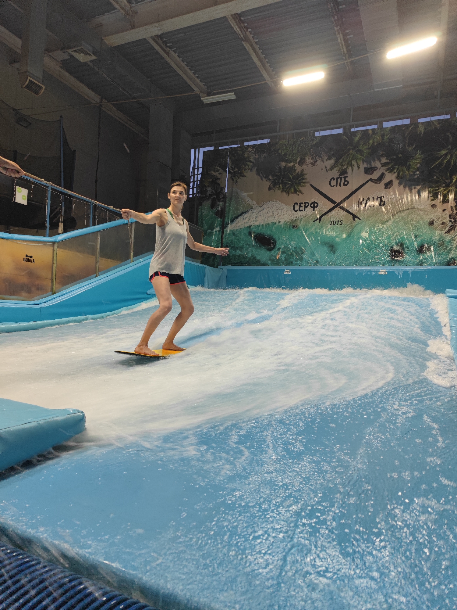 This media file captures a young woman indoor surfing on an artificial wave in a surf training center. The woman, wearing a grey tank top and black shorts with pink trim, is balancing on a yellow surfboard. She is mid-action, with her arms extended for balance as she rides the wave. The setting appears to be an indoor facility with a large blue pool and a ramp structure. The background features a large banner with palm trees, sand, and ocean imagery, along with text that appears to be in Russian, including "СПб" and "КЛУБ" and "2015". Another banner with the word "GORILLA" is visible on a yellow section of the pool wall. The lighting suggests it is daytime, though the exact time is not discernible. The overall mood is one of recreation and active pursuit.