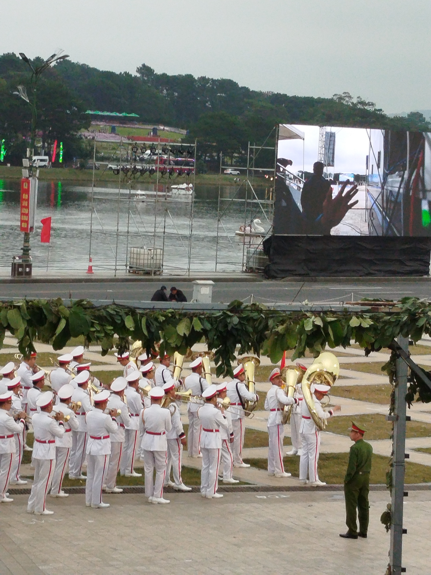 In Da Lat, Vietnam, a marching band in white uniforms with red trim is positioned in the foreground, playing brass instruments. A lone soldier in a green uniform stands to the right, facing away from the band. Behind them, a body of water is visible, bordered by scaffolding and a large screen displaying an image of a hand. Trees and buildings form the backdrop, with a cloudy sky overhead. The scene appears to be set in a public park or plaza, possibly during an event or ceremony.