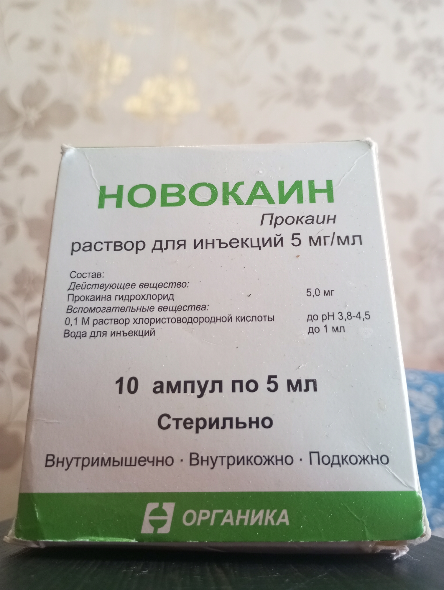A close-up, eye-level shot captures a white cardboard box for a pharmaceutical product. The box is positioned slightly tilted, with the top left corner closer to the camera. The background is a lightly textured wallpaper with a floral pattern in muted beige and cream tones.
The dominant text on the box is in large, bold green sans-serif font, reading "НОВОКАИН" (NOVOCAIN). Below this, in smaller black text, are the words "Прокаин" (Procaine) and "раствор для инъекций 5 мг/мл" (solution for injection 5 mg/ml).
Further down, a section labeled "Состав:" (Composition:) lists the contents. Under "Действующее вещество:" (Active substance:) is "Прокаина гидрохлорид" (Procaine hydrochloride). Under "Вспомогательные вещества:" (Excipients:) are "0,1 М раствор хлористоводородной кислоты" (0.1 M hydrochloric acid solution) and "Вода для инъекций" (Water for injections). To the right of these, smaller text indicates "5,0 мг" (5.0 mg) next to the active substance and "до рН 3,8-4,5" (to pH 3.8-4.5) and "до 1 мл" (up to 1 ml) next to the excipients.
At the bottom of the visible portion of the box, a line of text reads "10 ампул по 5 мл" (10 ampoules of 5 ml). Below this, in bold black text, is "Стерильно" (Sterile). The final line of text, in black sans-serif font, indicates administration routes: "Внутримышечно · Внутрикожно · Подкожно" (Intramuscularly · Intracutaneously · Subcutaneously).
At the very bottom of the image, a green band across the box features a white stylized logo and the text "ОРГАНИКА" (ORGANIKA). The box appears slightly worn, with minor creasing visible on the edges and surfaces. The lighting suggests an indoor environment, possibly a room with ambient light.
The provided image does not contain any elements or information that would allow for the determination of its location in the city of Kirzhach, Russia.