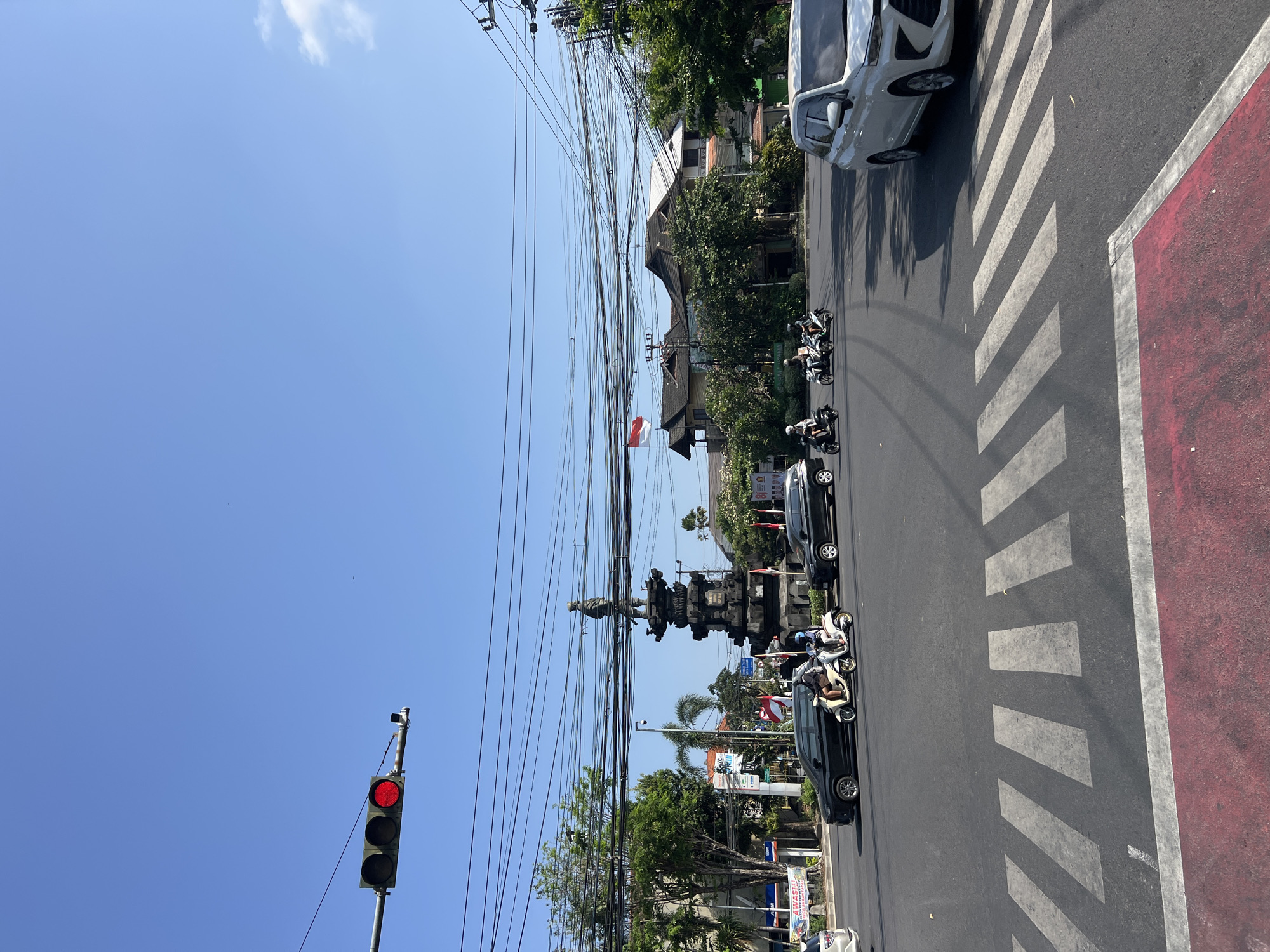 A wide-angle, eye-level shot captures a sunny day at a busy intersection in Denpasar, Indonesia. A traffic light at the top left shows a red signal. Dominating the center of the frame is a tall, dark stone monument topped with a statue of a person. The monument is surrounded by lush green trees and complex overhead wiring. The intersection features a pedestrian crosswalk with white stripes and a red border in the foreground. Several vehicles are present: a black car and a light-colored scooter with two occupants are moving through the intersection. Another black car is stopped at the intersection, and a white SUV is visible on the right side of the frame. Buildings with traditional architecture, including a prominent Indonesian flag, are visible in the background. The sky is a clear, bright blue, with a few wispy clouds on the right.