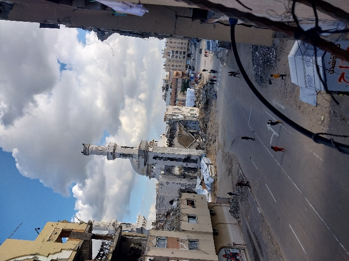 This image displays a street scene in Khan Yunis, Palestinian Territory, characterized by widespread structural damage to buildings under a partly cloudy sky.
In the mid-ground, a large mosque with a white dome and minaret stands, exhibiting significant damage to its exterior facade and surrounded by piles of rubble. To the left, a multi-story building is partially collapsed, revealing exposed floors and interior elements. On the right, a heavily damaged building's facade is visible, with white laundry hanging from an upper-level opening.
The paved road in the foreground features white lane markings, including a directional arrow. Several individuals are present on the street: one person in a red top walks near the center, another in dark clothing is to their right, and approximately ten additional people are observed walking further down the road, some gathered near the damaged mosque and others in the distant background. A person in a yellow top is also visible walking on the right side of the road.
A white tent-like structure, bearing partial Arabic text, is visible in the lower right foreground, partly obscured by a dark, curved cable or wire. A graphic resembling the character Deadpool is painted on the wall of a damaged building on the left side of the street. Vehicles are present in the distant background.