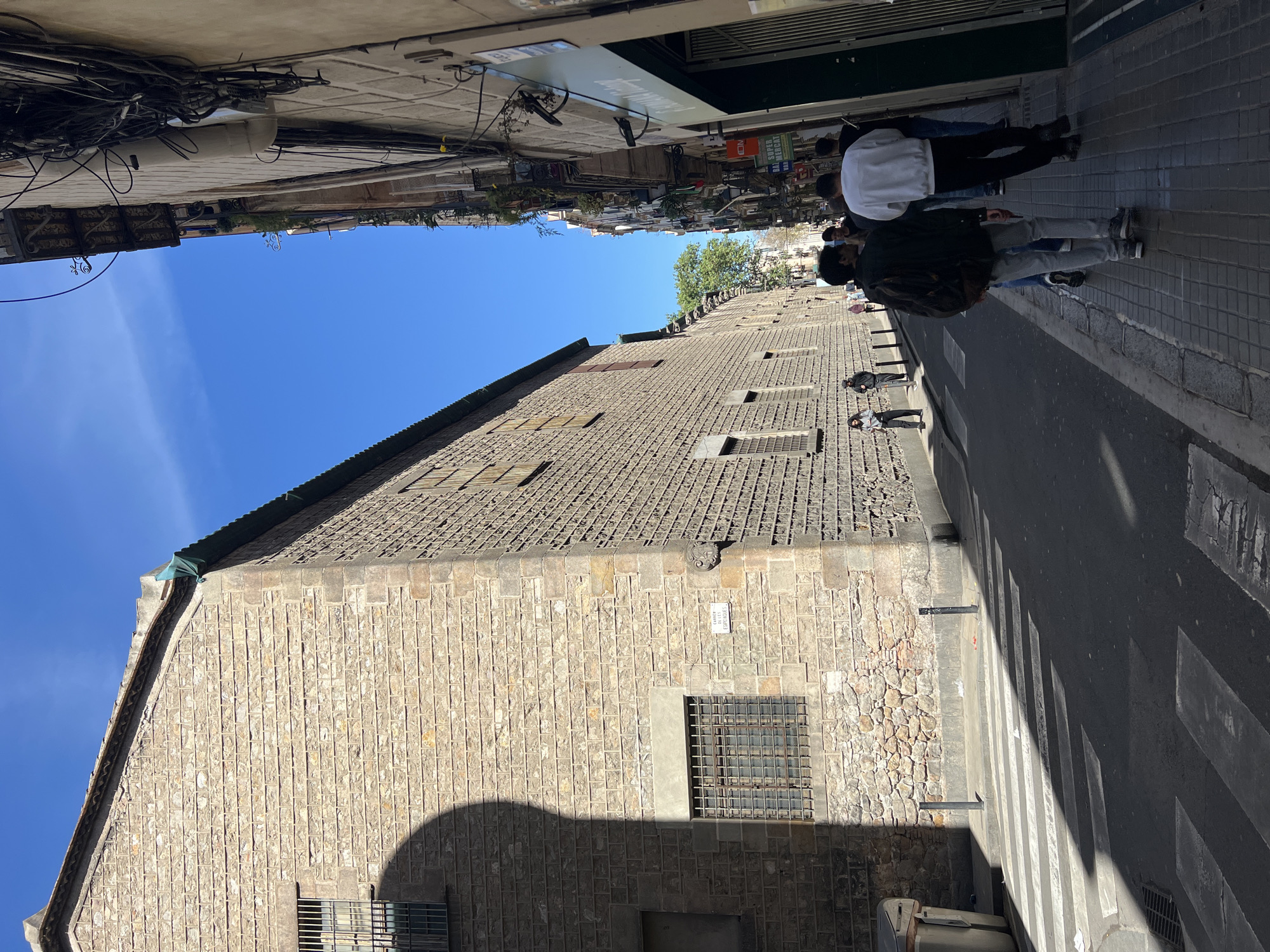 The image depicts a vibrant street scene in Barcelona, Spain, specifically within the historic Barri Gòtic, under a clear, bright blue sky. The perspective looks down a narrow urban street, with a prominent stone building dominating the left side and a paved sidewalk and road on the right.
On the left, a large, old stone building rises, featuring a textured facade of light-colored blocks and a sloped, tiled roof. Several rectangular grated windows are visible on its side. A white street sign is affixed to this building, clearly displaying "CARRER DE / CALDERO / N. 21-25 / BARRI GÒTIC," confirming the location as Barcelona's Gothic Quarter. The building casts a long shadow across the street and sidewalk, indicating the sun is high, likely late morning or early afternoon.
On the right side of the street, a paved sidewalk runs parallel to the road. Numerous thick black electrical wires and conduits are tangled overhead, attaching to the buildings. Several pedestrians are visible on the sidewalk. In the middle ground, three individuals walk together: one wearing a white jacket, another in a dark jacket and grey pants, and a third partially obscured. Further down the street, more people are present, some standing near storefronts.
The road features a distinct pedestrian crossing marked with white zebra stripes. In the distance, other buildings line the street, with a glimpse of a green-leafed tree and other urban structures. A distant sign appears to read "SÚPER MERCAT." The overall atmosphere is that of a lively urban setting on a sunny day.