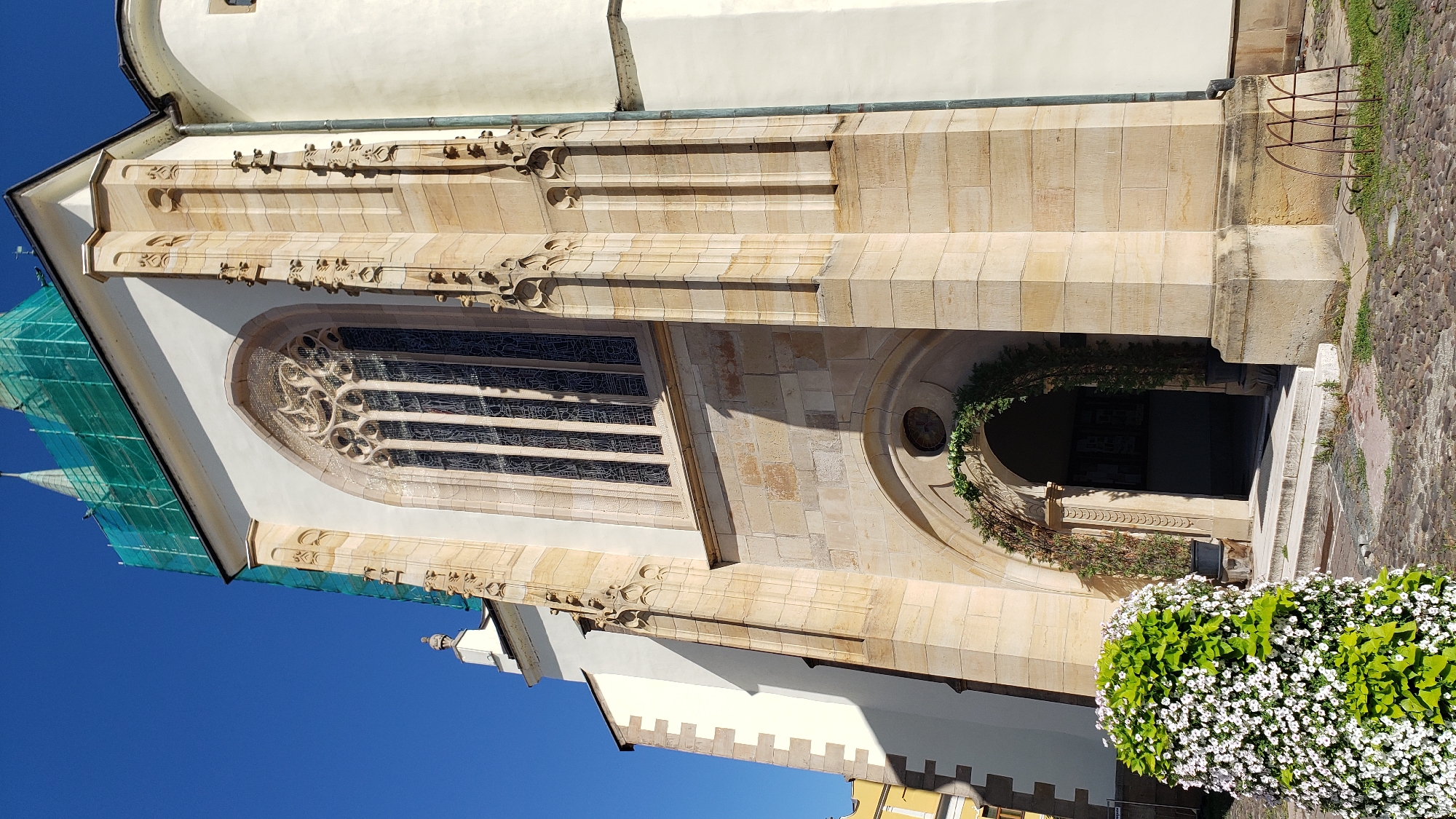 The image shows the exterior of St. Nicholas Cathedral in Prešov, Slovakia. The facade is made of stone and features a large Gothic window with intricate tracery. The entrance is an arched doorway adorned with greenery. In the foreground, a large bush with white flowers is visible. The sky is clear and blue. Scaffolding is present on the upper portion of the building, indicating ongoing renovations or construction.