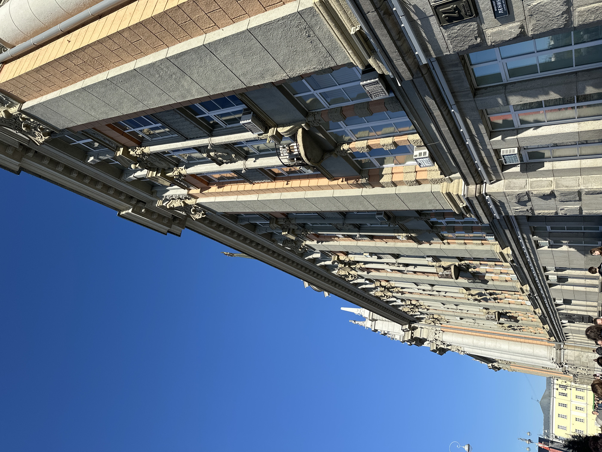 The image shows a building facade in Yekaterinburg, Russia, against a clear blue sky. The building is of classical design, with multiple stories and features detailed stonework, windows with air conditioning units, and decorative architectural elements like cornices and columns. Some people are visible on the right side of the image, but they are indistinct. The building seems to be a significant architectural landmark. Markings in Cyrillic script are visible on the building's exterior.