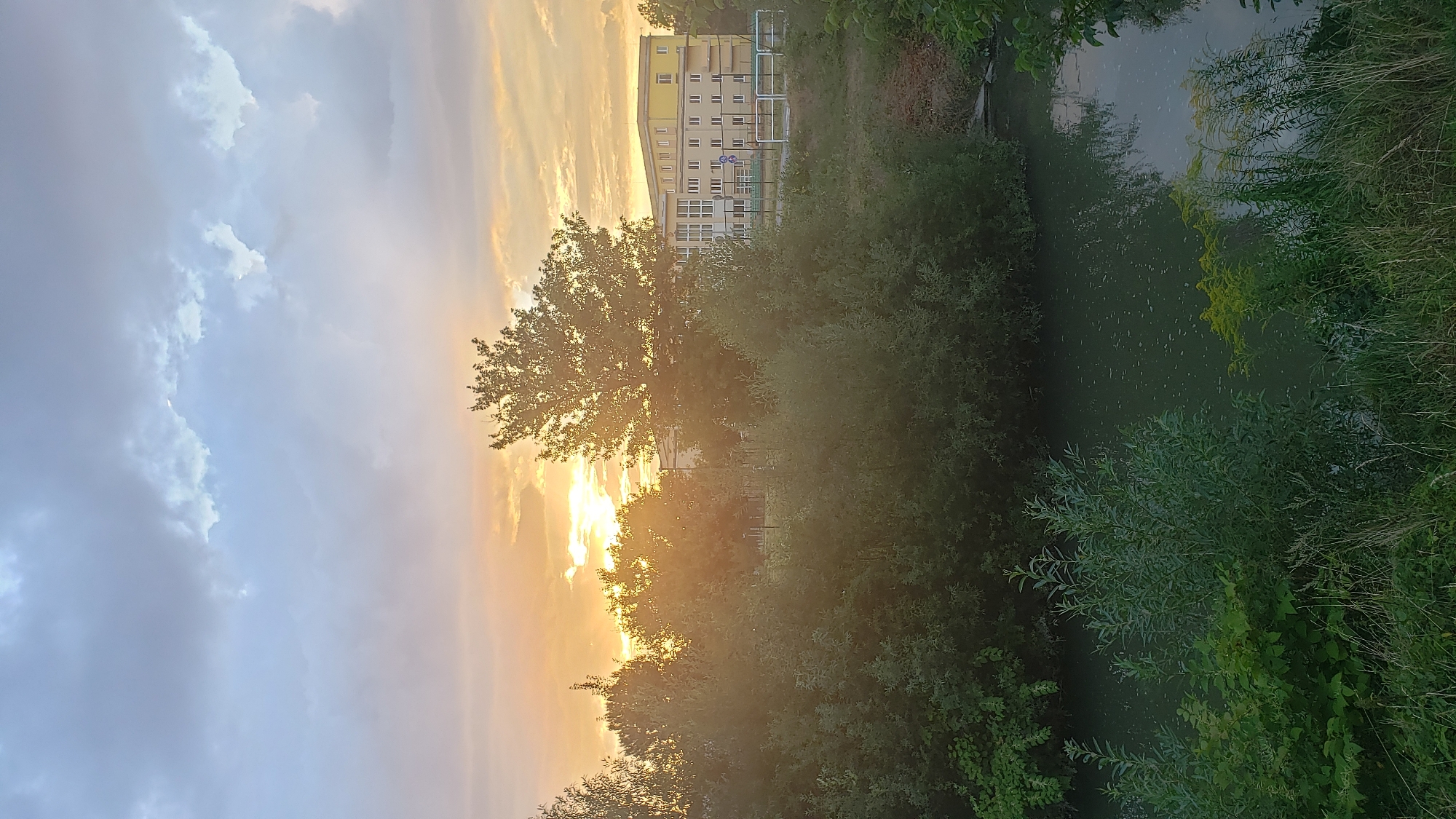 The image depicts a serene waterscape with a building in the background, set against a twilight sky. The foreground features a body of dark water, possibly a river or canal, lined with lush green vegetation. Trees and bushes dominate the midground, with some appearing to be on the bank of the water.
In the distance, a pale yellow, multi-story building stands prominently. It has a rectangular structure with numerous windows and appears to be a modern complex, possibly an educational institution or residential area. The building is partially obscured by the trees.
The sky above is a mix of dramatic clouds, with patches of orange and yellow light suggesting either a sunset or sunrise. The sun itself is not directly visible, but its light is filtering through the clouds, casting a warm glow on the scene. The overall atmosphere is peaceful and natural, with a hint of urban presence from the building.
While the image does not provide explicit clues to pinpoint a precise location within Prešov, Slovakia, the presence of a substantial building adjacent to a waterway, surrounded by greenery, is characteristic of many urban or suburban areas. It could be near a park, a university campus, or a residential district that incorporates natural elements. Without further context or distinctive landmarks, specifying the exact location within Prešov is not possible.