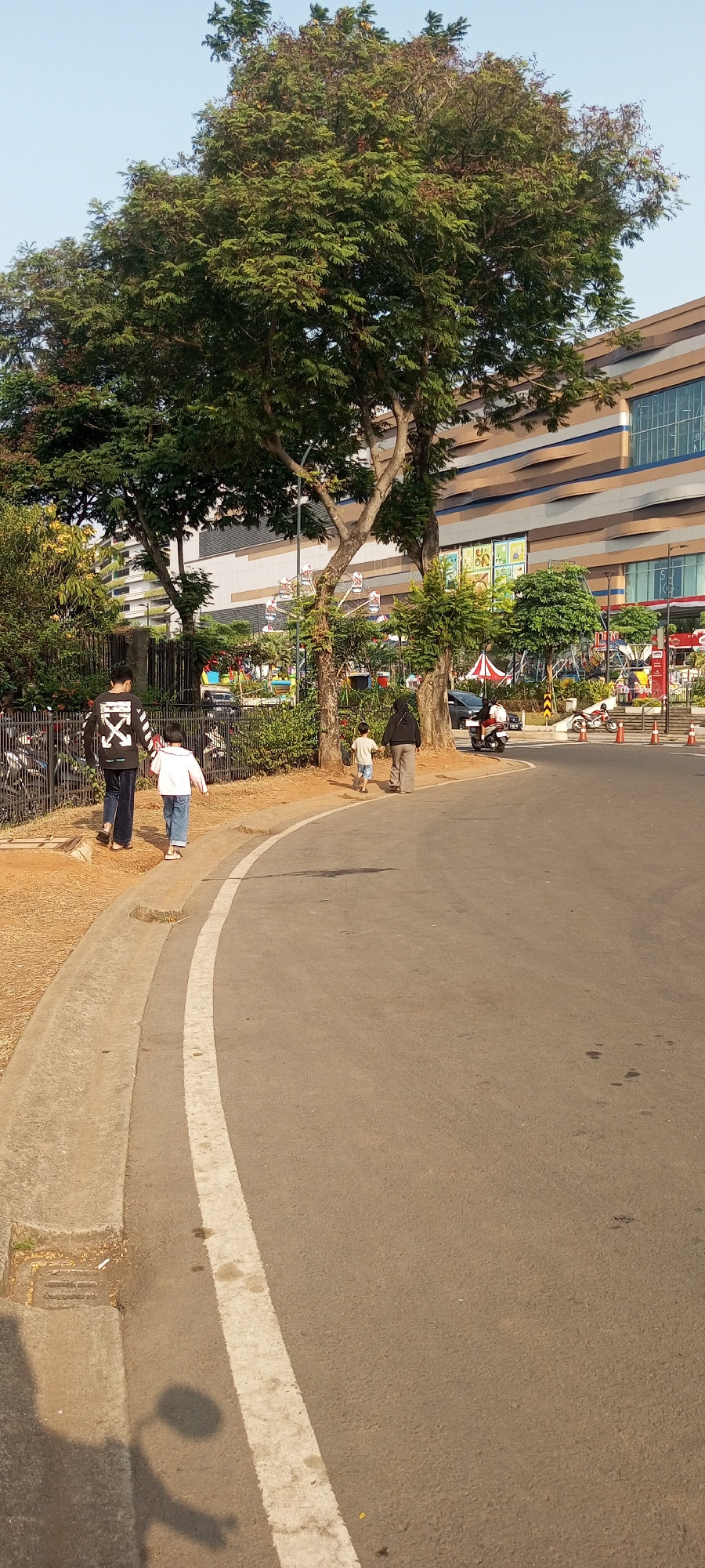 The image depicts a street scene in Lambangjaya, Indonesia, with a large building and a Ferris wheel visible in the background. In the foreground, four individuals are walking along the side of a paved road with a white line marking its edge. Two adults and two children are present, walking in the same direction. One adult, wearing a black long-sleeved shirt with a white graphic, walks with a child in a pink jacket and jeans. Further ahead, another adult in dark clothing walks with a child in a light-colored shirt and shorts. The road curves to the right, and traffic cones are placed along the edge of the road further down. The environment includes trees, a metal fence, and parked motorcycles on the left side of the road. A shadow of a motorcycle and rider is cast on the road surface. The sky is clear and blue.