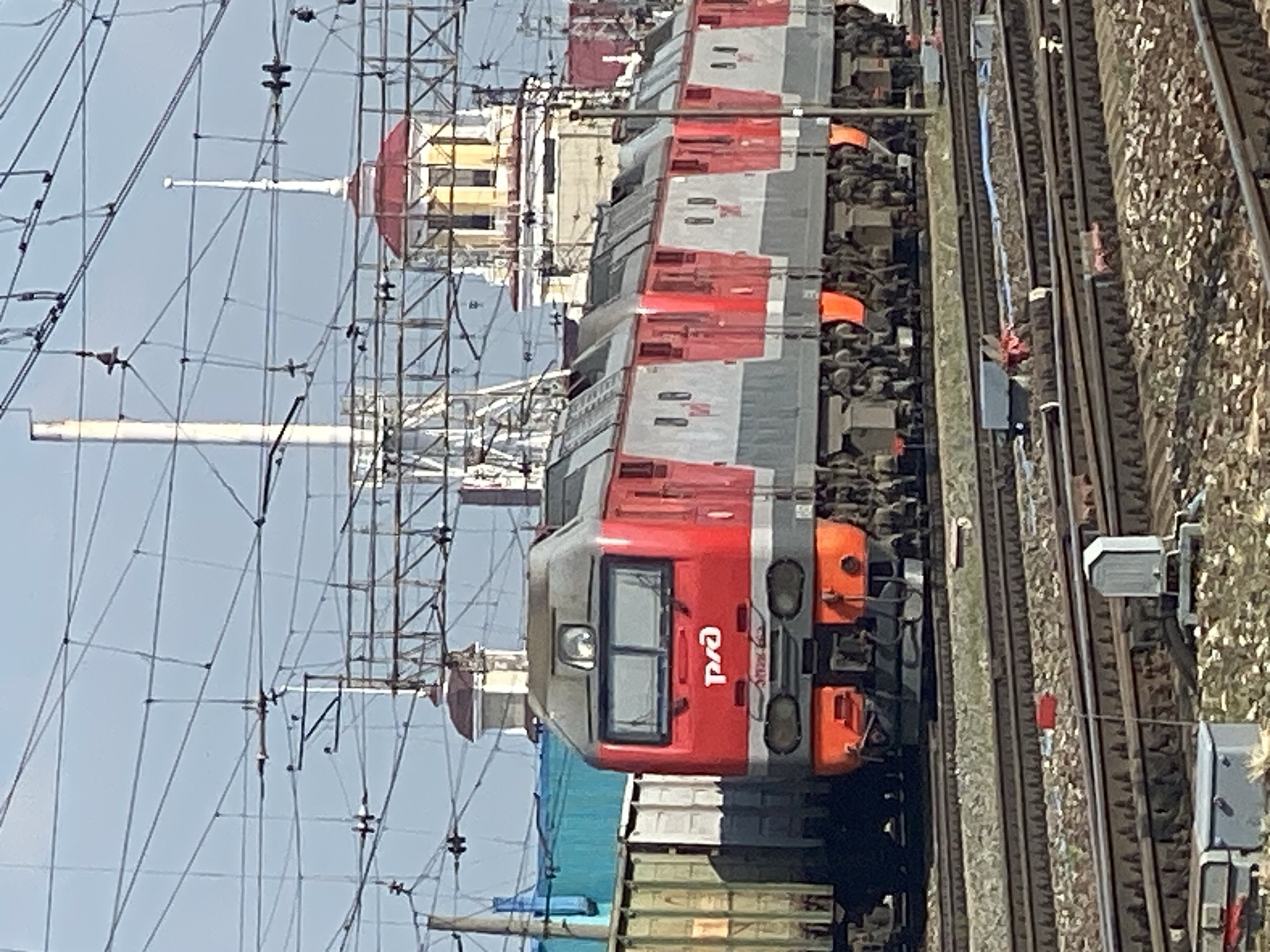 A red and gray electric locomotive, labeled "РЖД", is stationary on train tracks in Novokuznetsk, Russia. The locomotive is a multi-unit train, with at least six cars visible, each featuring red and gray paneling with small windows. In the background, there are industrial structures, including buildings with distinctive red roofs and yellow accents, as well as numerous overhead power lines and supporting poles. To the left of the locomotive, a section of a green freight car is visible. The foreground shows more train tracks and ballast, with some small objects and industrial equipment scattered near the tracks. The sky is clear and blue. The image appears to have been taken during daylight hours.