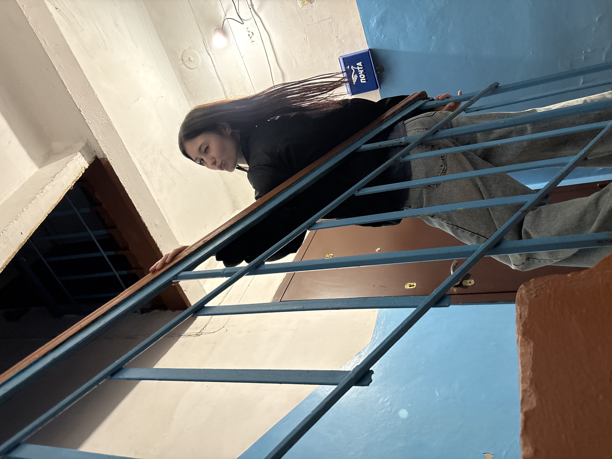 A young woman with long, dark hair, wearing a black jacket and light-colored jeans, is leaning over a blue metal railing in what appears to be an indoor stairwell or hallway. The image is captured from a low angle and is rotated, making the scene appear tilted. Her hair flows downwards (relative to the image frame) over the railing. She holds onto the wooden top of the railing with her right hand, which is visible. Her expression is a slight pout, with her lips pursed.
The setting is a building interior, with walls painted in contrasting white and light blue. Above and behind her head, a bare light bulb illuminates the area. To her right, mounted on the blue wall, is a blue mailbox with the white Cyrillic text "ПОЧТА" (Pochta), meaning "Mail" or "Post Office." In the background, past the railing and her legs, a brown door with golden hardware is partially visible, showing what appears to be the number '5' on it. The overall impression is that of an older, possibly residential, building in Фоки, Russia.