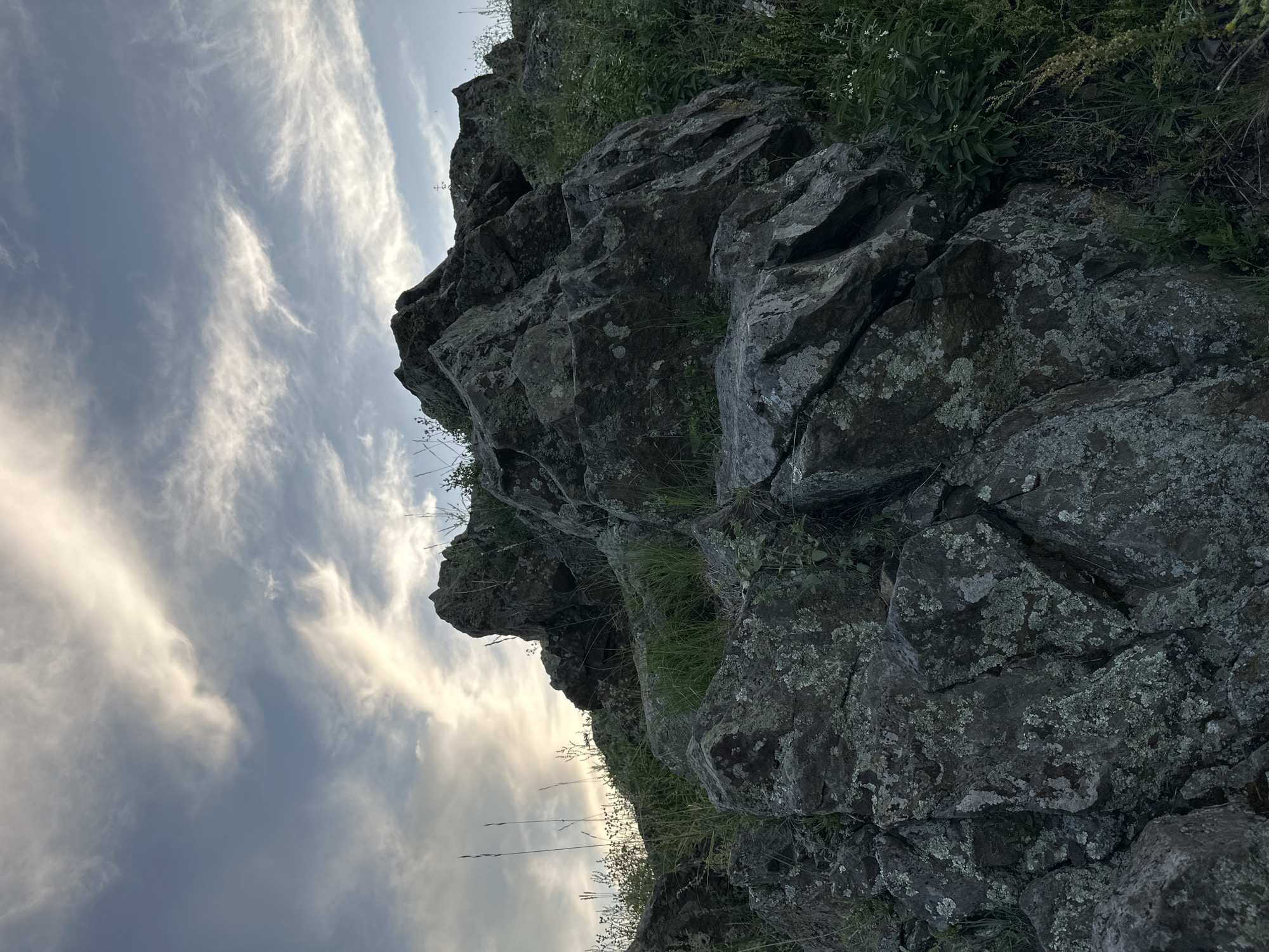 This media file features a close-up, low-angle shot of a rocky cliff face with vegetation growing on it, set against a cloudy sky. The rocks are dark gray and appear weathered, with patches of lichen clinging to their surfaces. Tufts of green grass and other plants sprout from crevices in the rocks and along the top edge of the cliff. The sky above is filled with wispy, white clouds against a pale blue background, suggesting either early morning or late afternoon light. The overall impression is one of a natural, rugged landscape. There is no text visible in the image.