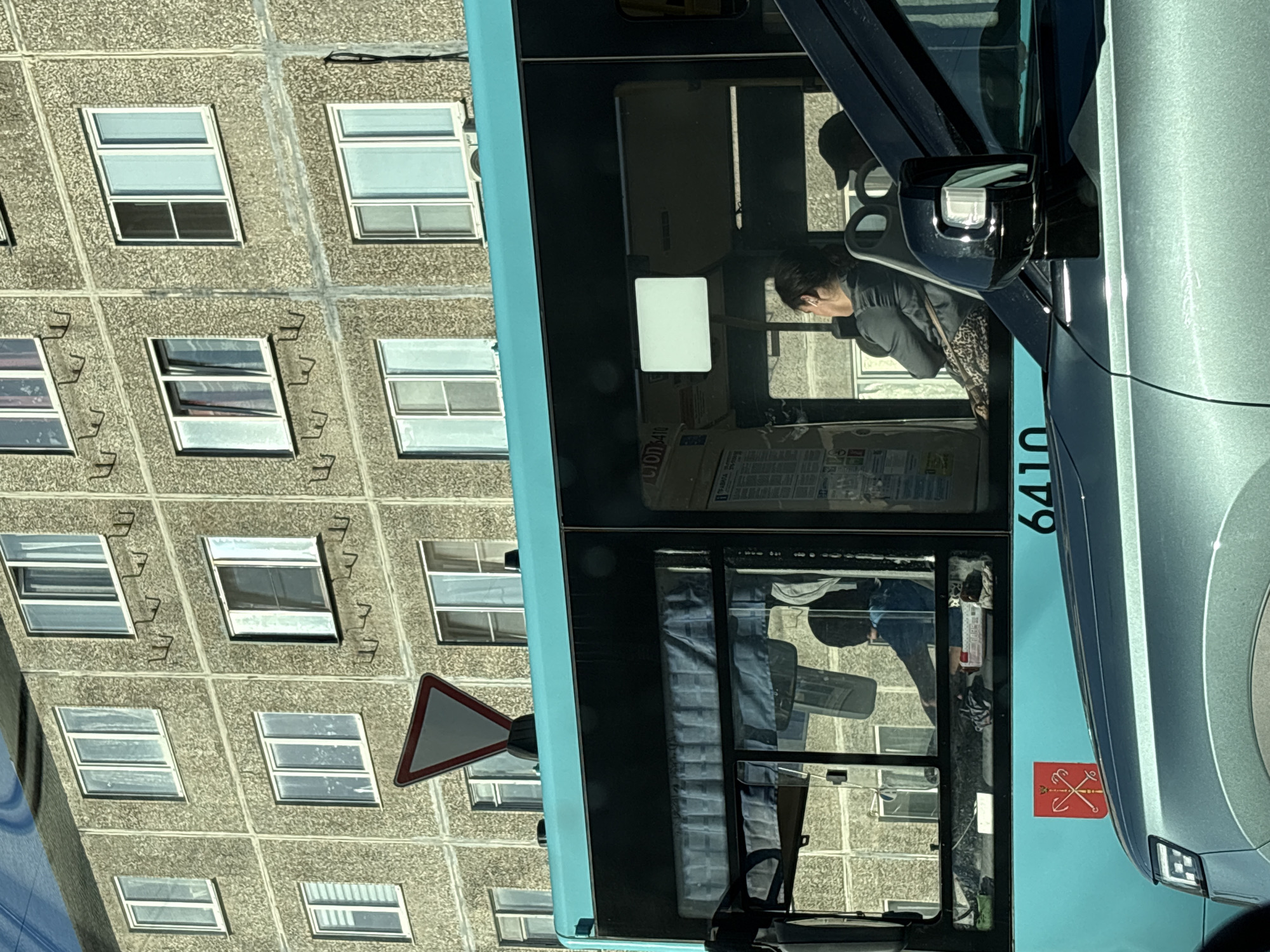 The image displays an urban street scene in Saint Petersburg, Russia, identified by the distinctive coat of arms on a public transit bus. In the foreground, the front right quarter of a dark teal or grey car is visible, including its headlight and side mirror. The midground features a teal-colored public transit bus, prominently displaying the red and white coat of arms of Saint Petersburg (a crossed scepter and anchor) on its side, above the fleet number "6410". A red and white triangular "Give Way" road sign is mounted on the bus's front roof. Inside the bus, a female passenger with dark hair pulled back is seated, looking towards the right, wearing a dark shirt and a patterned skirt or pants. Reflected in the bus window, the silhouette of another individual, possibly the driver, wearing a dark cap, is visible. A rectangular white screen or light is mounted inside the bus above the passenger. The background consists of a multi-story, grey, textured concrete residential building with numerous white-framed windows, some of which are open. Metal fixtures, possibly hooks, are regularly spaced on the building's facade, and an air conditioning unit is attached below a window on the building's right side. The scene is illuminated by natural daylight.