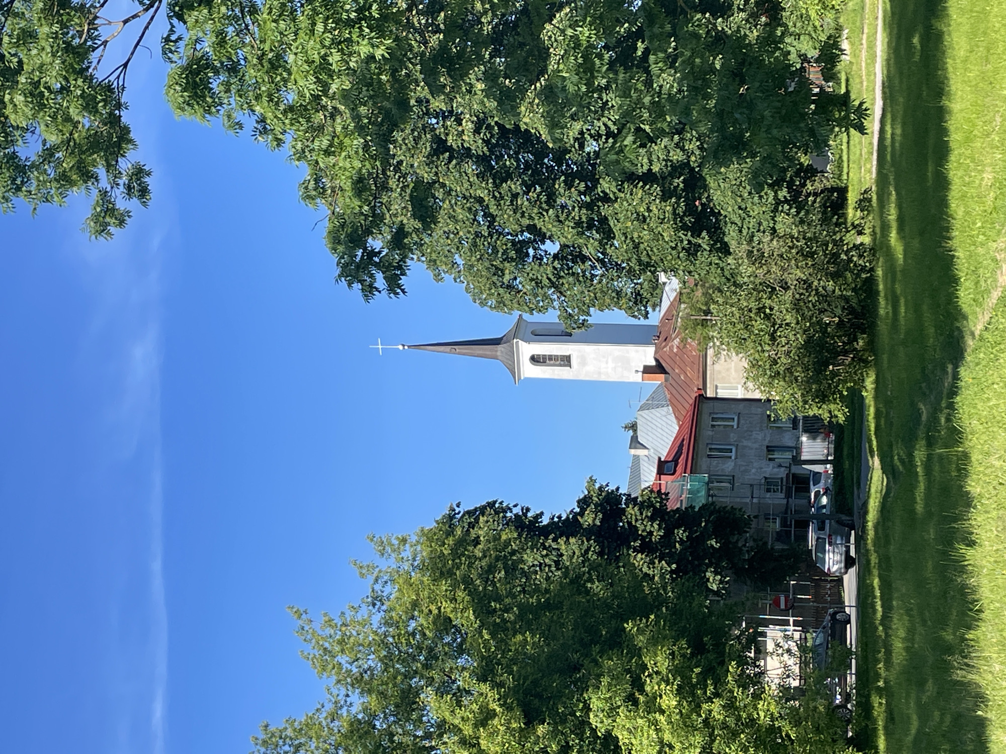 This image depicts a view of a church tower and surrounding buildings, partially obscured by lush green trees. The sky is a clear, vibrant blue, indicating fair weather during daylight hours. The orientation of the photo suggests it might have been taken vertically and then rotated for display.
The central subject is a white church tower with a dark, pointed spire topped by a cross. Adjacent to the church are residential or commercial buildings with red-tiled roofs and grey facades. Some of the buildings appear to be undergoing renovation, with scaffolding visible. A few cars are parked along a street in front of the buildings.
The foreground and midground are dominated by the dense foliage of trees, creating a natural frame for the architectural elements. The background features a well-maintained grassy area, possibly a park or lawn, with shadows cast by the trees, hinting at the time of day being late morning or early afternoon.
The overall scene evokes a peaceful, summery atmosphere in an urban or semi-urban setting within Tallinn, Estonia. No people are clearly visible, and no text is discernible in the image.