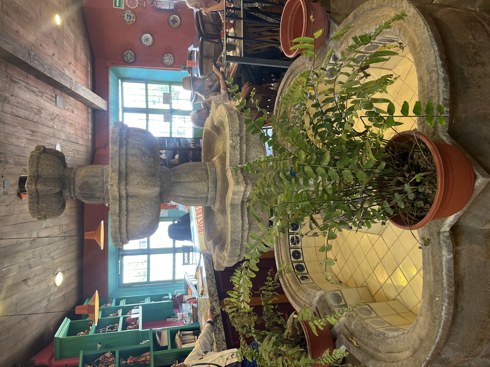 Indoor shot of a two-tiered stone water fountain in a restaurant in Puebla, Mexico. The water basin on the bottom is tiled with small diamonds. There are potted ferns sitting on top of both tiers of the fountain. People can be seen in the background eating at tables. The interior of the building has a rustic design with wooden walls and an exposed beam ceiling. There are windows behind the fountain.