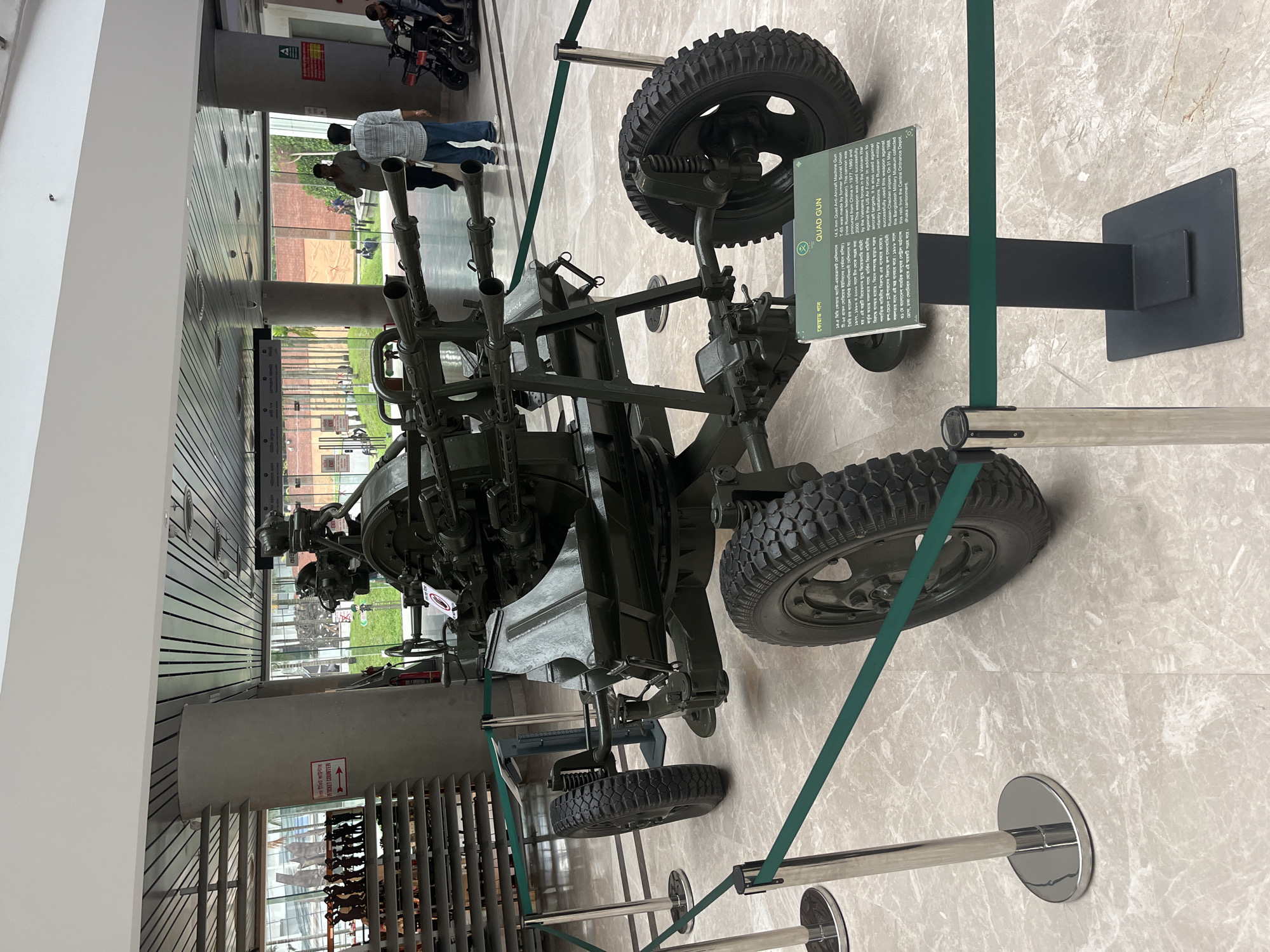 The image shows a Quad Gun, specifically a 14.5mm Quad Anti-Aircraft Machine Gun T-65, exhibited in what appears to be a museum. This weapon was made by the former Soviet Union and procured from China. According to the signage, the museum acquired this relic from the Central Ordnance Depot, Dhaka Cantonment, on May 31, 1988. The weapon is behind a barrier of metal poles and green fabric strips. The flooring is a beige marble pattern. In the background, partially visible through a glass wall, are people, trees, and potentially other exhibits.
