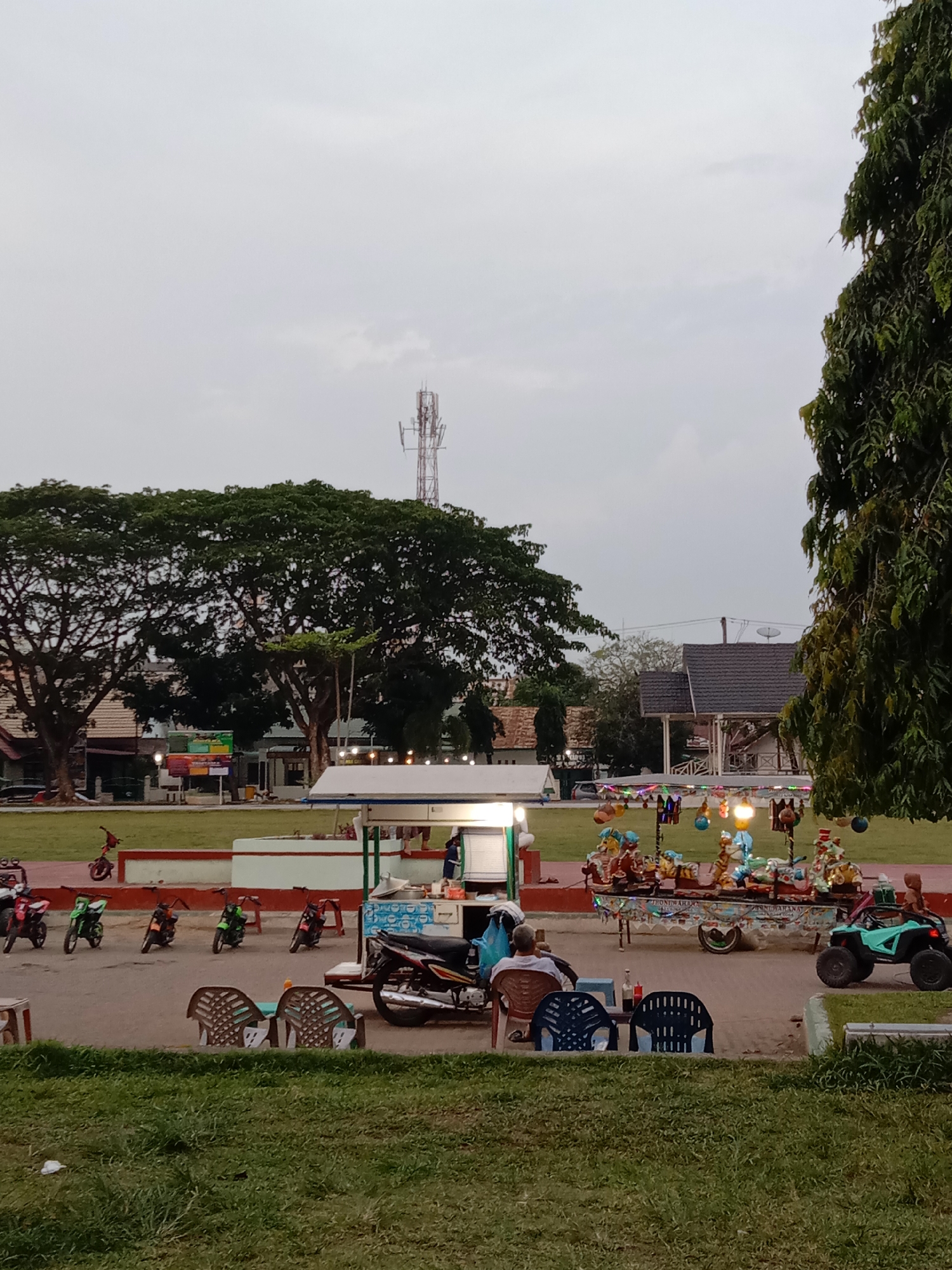 This media file depicts an outdoor public space, likely a park or town square, in Lhokseumawe, Indonesia, captured during dusk or on an overcast day. A wide expanse of green grass dominates the foreground and mid-distance, bordered by large, mature trees. In the background, traditional buildings with dark roofs are visible, and a prominent telecommunications tower stands against the cloudy sky.
The central activity occurs on a paved area running through the space. Here, a brightly illuminated vendor stall, possibly selling food or beverages, draws attention. Nearby, a colorfully decorated cart, adorned with numerous lights, animal figures, and other ornaments, suggests a mobile toy or snack vendor, accompanied by a small turquoise ATV, possibly a children's ride. Several plastic chairs and small tables are set up around the vendor stall, with at least two individuals seated and relaxing, one with their back to the camera. A collection of motorcycles, including several small, brightly colored children's ride-on bikes, are parked in the vicinity.
The scene conveys a relaxed, casual atmosphere, typical of a community gathering spot for leisure and family recreation. Artificial lights from the stalls and carts illuminate the area, contrasting with the soft, ambient light of the sky. Visible text on the decorated cart includes partial phrases such as "...IHONG DARAHA..." and "...JINNI TARIANA...", possibly brand names or design elements. On the side of the main vendor stall, a blue label with fragmented text, possibly "COOK..." and "FRESH DRINK," is discernible.