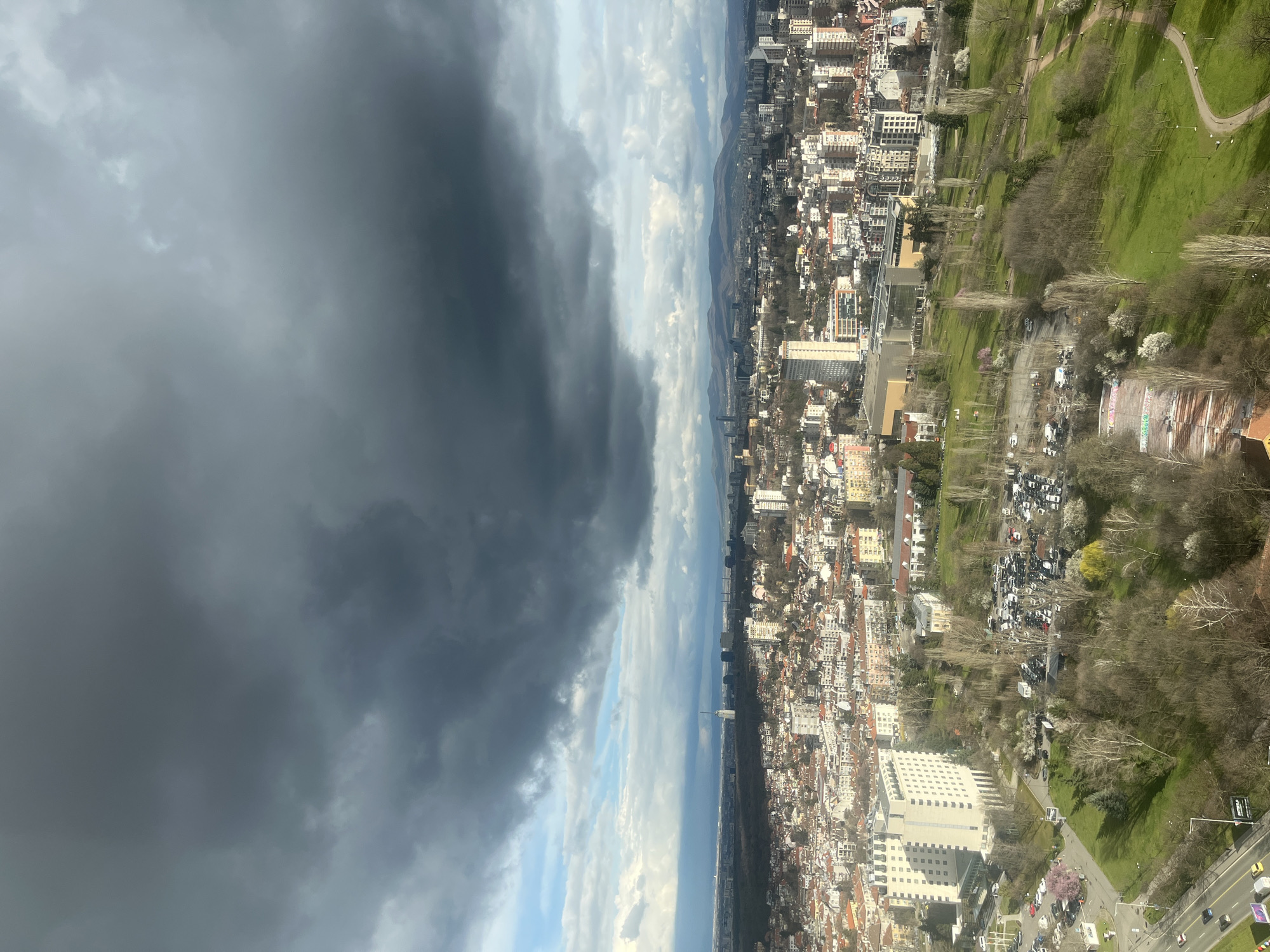The image shows an aerial view of Sofia, Bulgaria, with a distinct contrast between a dark, stormy cloud covering the left side and the city on the right. The city consists of closely packed buildings, ranging from low-rise residential structures to taller apartment complexes. A green space, likely a park, borders the city with trees and paths visible. A street with some cars can be seen alongside the park. The overall environment is urban, with signs of nature integrated into the landscape.
