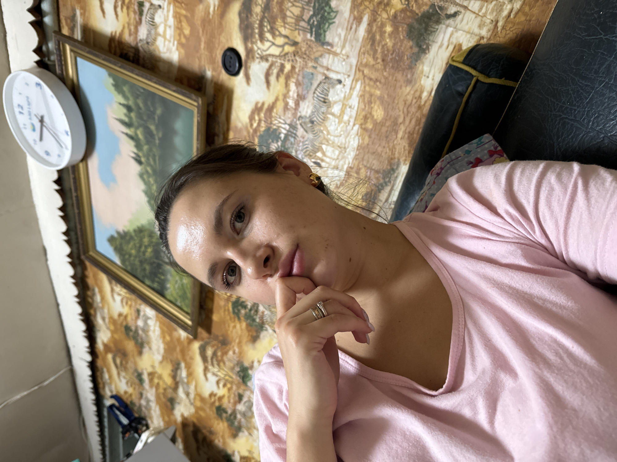 A woman wearing a pink shirt is resting her chin on her hand. She has brown hair and a ring on her finger. Behind her is a wall decorated with a tapestry depicting animals. There's a picture frame on the wall, and a round clock. The environment appears to be inside a home, although the specific location in Красный Маяк, Russia, cannot be determined from the image.