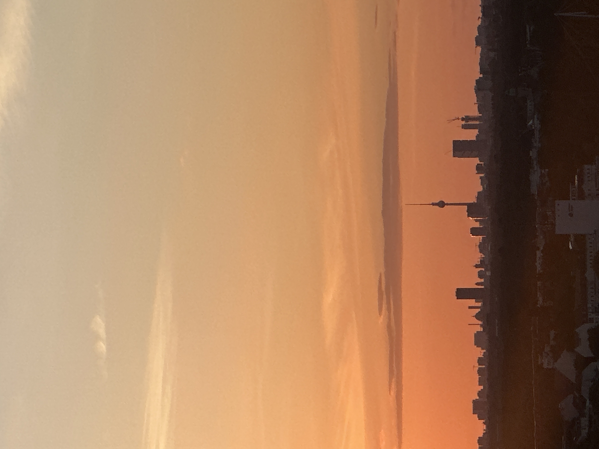 The image shows a skyline of Berlin, Germany during a sunset. The sky is a gradient of orange and yellow, with some thin clouds. In the foreground, there is a dark silhouette of trees and buildings. The prominent features of the Berlin skyline include the Fernsehturm (TV Tower) on the left, a tall, dark rectangular skyscraper, and other buildings of varying heights. The sunset casts a warm glow over the city.