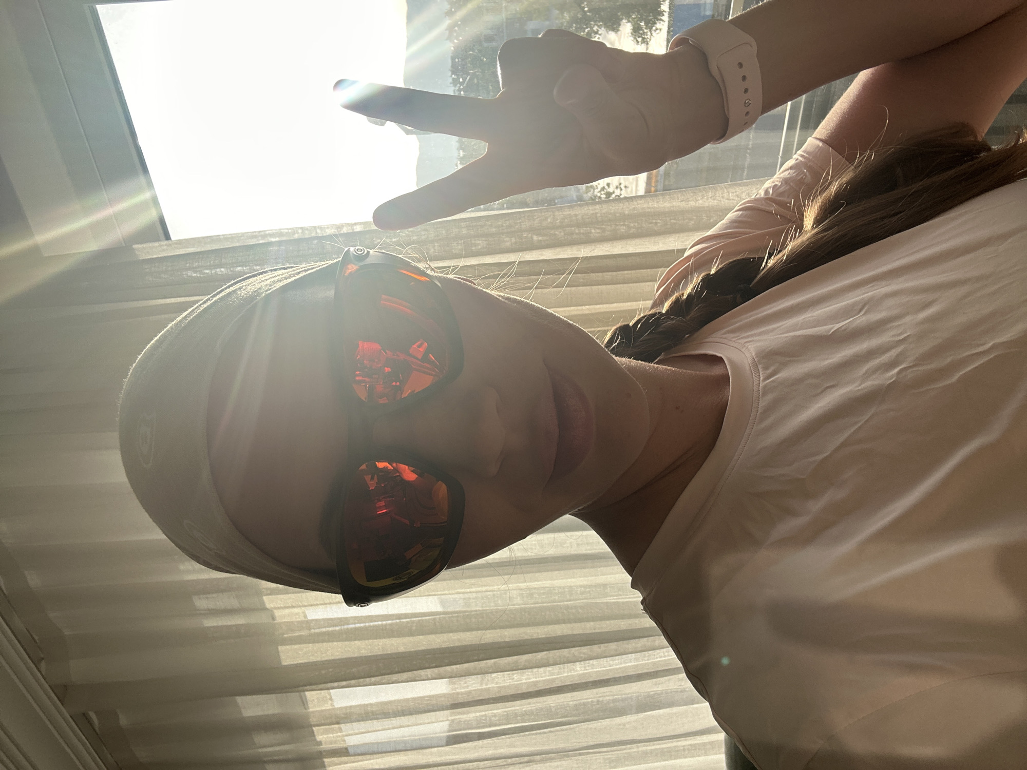 A person is standing in front of a window with closed blinds. The person is wearing a light pink t-shirt, sunglasses, and a headband, and has a braid in their hair. They are making a peace sign with their hand. The person's wrist has a pink fitness tracker. The sunglasses have red lenses that reflect the environment, but the location and reflections are not specific enough to determine if it is in Fethiye, Turkey. The location could not be determined based on this photo.