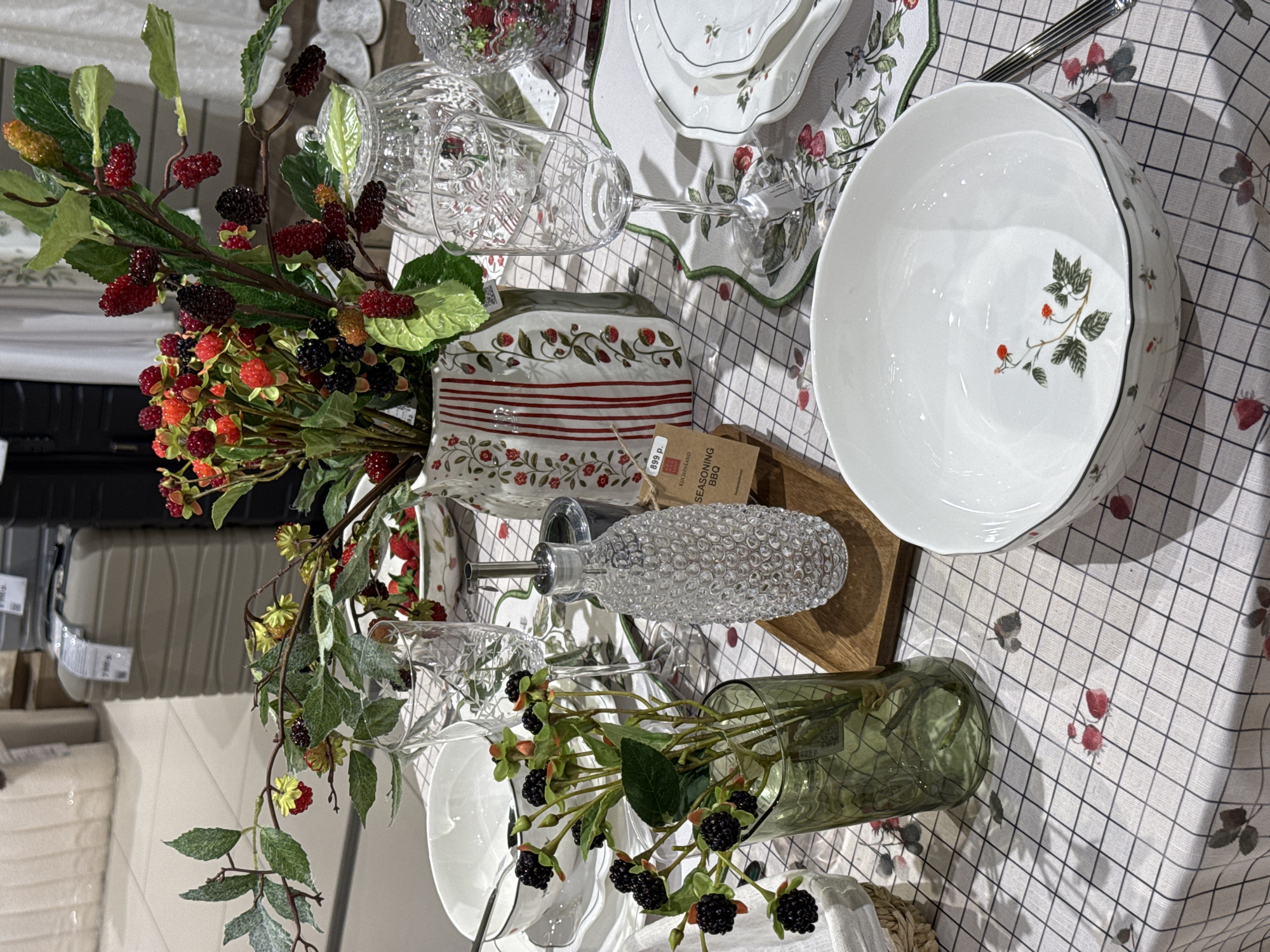 This image depicts a detailed retail display of tableware and home decor, situated within a Kuchenland Home store in Perm, Russia. The foreground features a table covered by a light-colored tablecloth with a subtle black grid pattern and scattered illustrations of red berries and green leaves.
On the table, a formal place setting is arranged. This includes a large white ceramic serving bowl with a wavy rim, thin black border, and internal red berry and leaf motifs, positioned in the lower right. To the left, a stack of white plates with a thin black rim and small berry accents is visible, with a silver spoon resting on the top plate. On the right, a white placemat with a green embroidered border and berry illustrations supports a stack of white plates with green borders and similar berry designs.
Glassware on display includes two clear stemmed wine glasses with etched geometric patterns and a large, clear, lidded glass jar, likely a candy dish, containing artificial red berries.
Two artificial berry arrangements serve as centerpieces. A dark green glass vase on the left holds branches adorned with dark green leaves and clusters of black berries. Central to the display is a white ceramic pitcher-style vase with vertical red stripes and climbing strawberry vine patterns, containing a prominent arrangement of artificial branches featuring a mixture of red, yellow-green, and black berries among green leaves. A smaller white bowl to the left of this vase contains loose artificial red berries.
A clear glass bottle with a textured, bobbled surface and a silver pour spout, likely an oil or vinegar dispenser, is placed on a small rectangular wooden tray. Attached to it is a brown tag indicating "899 p." (rubles), "KUCHENLAND HOME," and "SEASONING BBQ." A silver utensil is partially visible on the far right.
The out-of-focus background shows typical retail environment elements: stacked light-colored textiles on shelves in the upper left, and to their right, a light gray ribbed suitcase with a price tag, along with a darker suitcase, suggesting a section for travel accessories within the store. The floor is tiled with light-colored squares. No individuals are visible in the scene.