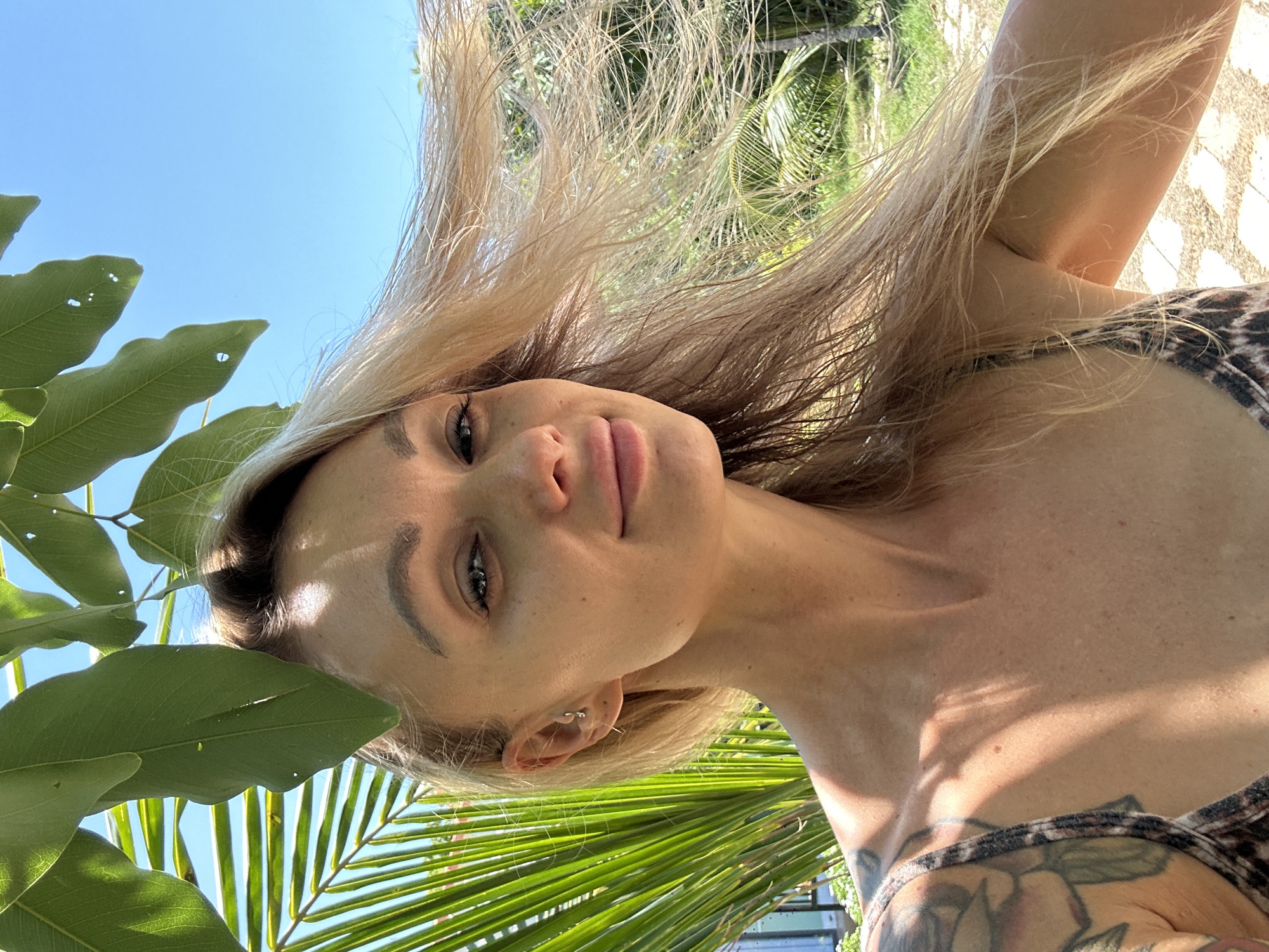 A woman with blonde hair is posing for a selfie in Mũi Né, Vietnam. The background includes lush green foliage, palm trees, and a clear blue sky. The woman is wearing an animal print top and has tattoos on her shoulder. She is smiling slightly.