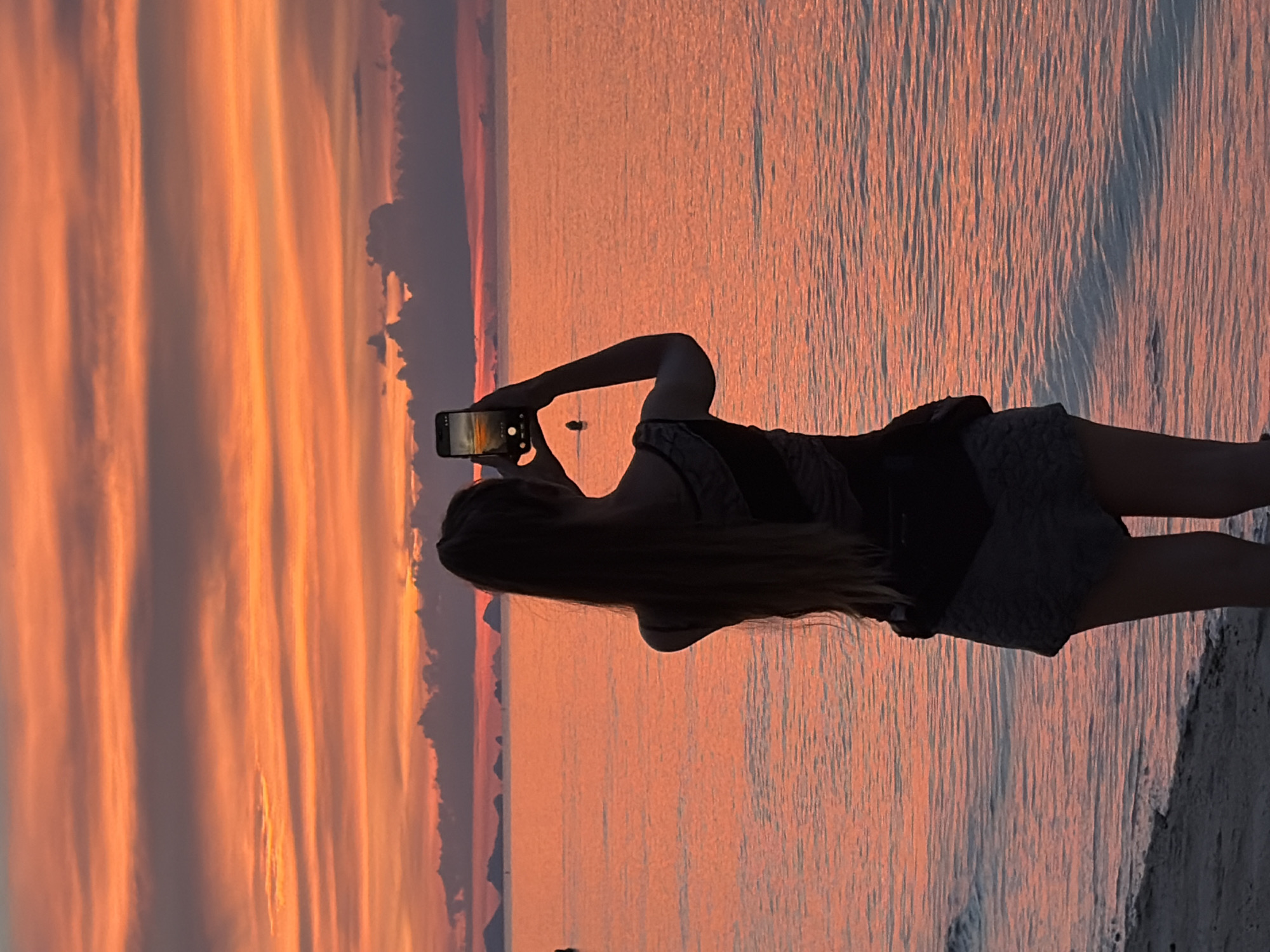 A woman is standing on a beach at sunset, facing away from the camera and towards the ocean, holding a smartphone up to take a photo of the sunset. The sky is a vibrant orange and pink, with layers of clouds. The ocean is calm and reflects the colors of the sky. In the distance, silhouetted islands are visible on the horizon. The woman has long, dark hair that is pulled back in a ponytail, and she is wearing a dark top and patterned shorts. The beach is sandy, and the waves are gently lapping at the shore. This scene is located in Baan Thong Po, Thailand.