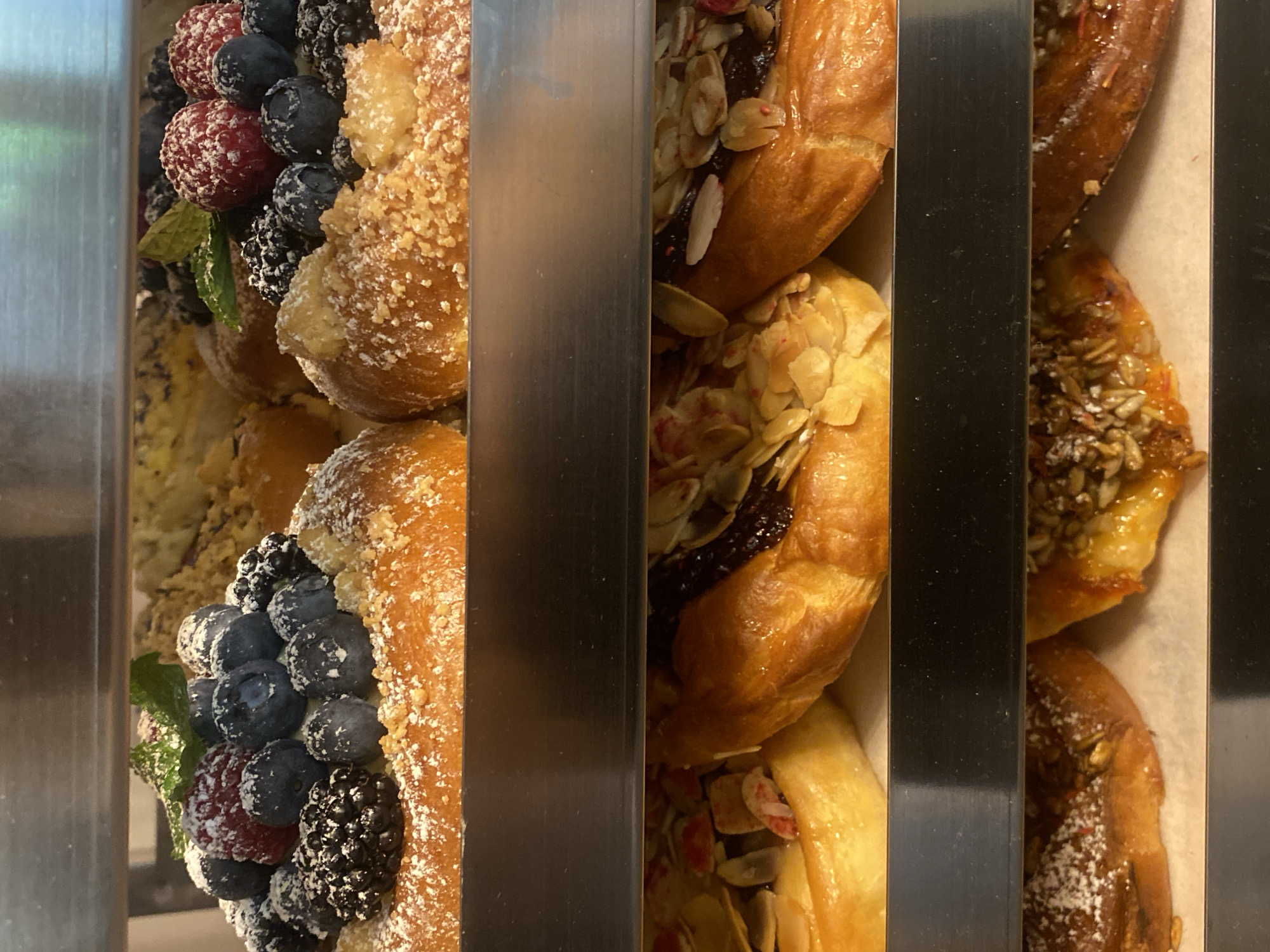 The image shows a close-up, vertical view of several pastries displayed in a bakery. The pastries appear to be on a shelf or in a display case, with metal dividers separating them into sections.
The leftmost section features a pastry topped with a generous assortment of fresh berries, including blueberries, raspberries, and blackberries. A few green mint leaves are also visible amongst the fruit. The pastry itself has a crumbly, textured topping and appears to be dusted with powdered sugar.
The next section displays pastries with a glistening jam filling, possibly fruit-based, and a topping of sliced almonds. The pastry has a golden-brown, flaky appearance.
Further to the right, another pastry is visible, topped with what looks like seeds, possibly sunflower seeds or a seed mix, and appears to have a glaze. This pastry also shows hints of powdered sugar.
The overall impression is one of a freshly prepared, attractive selection of sweet baked goods. The lighting suggests an indoor setting, likely a bakery. No people are visible in the image. The context provided indicates the location is Warsaw, Poland. There is no visible text in the image.