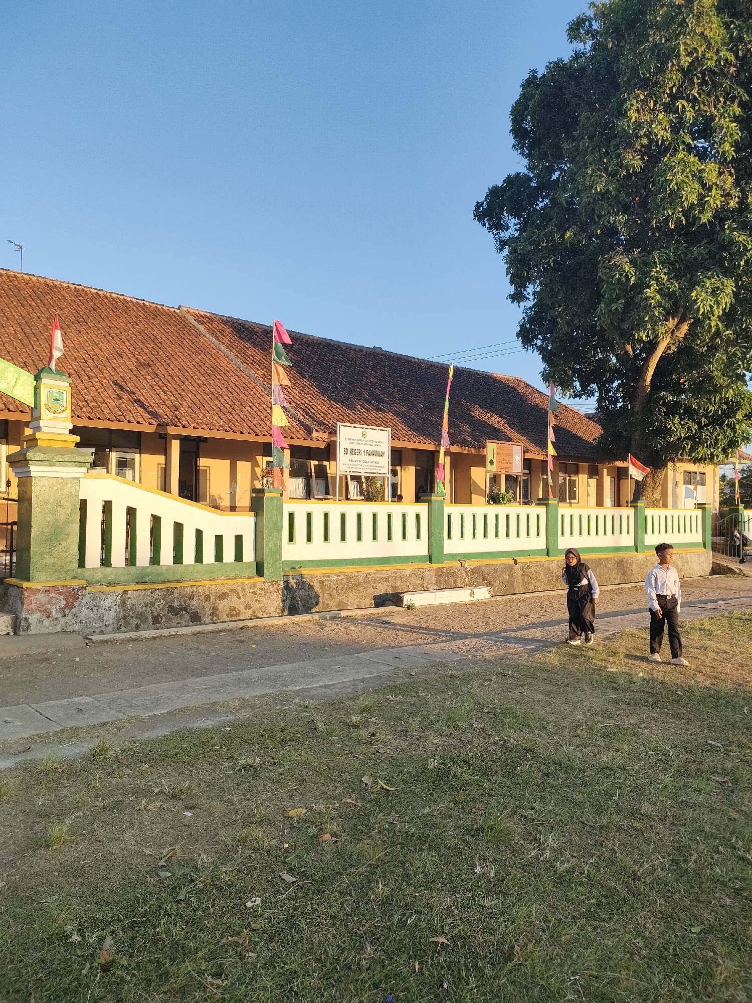 The image shows the facade of a school building under a clear blue sky. The building has a tiled roof and a yellow-painted exterior with several windows. A white fence with green accents runs along the front of the property. Two children, a boy in a white shirt and dark pants, and a girl in a dark hijab and pants, are standing on the sidewalk in front of the fence. A large tree with green foliage is visible to the right of the building. There are several flags planted in front of the school. A sign on the building reads "SD NEGERI 1 PANAWUAN". The ground in front of the school is a mix of pavement and grassy areas. The setting appears to be a rural or semi-urban area in Bojong, Indonesia. The lighting suggests it is late afternoon.