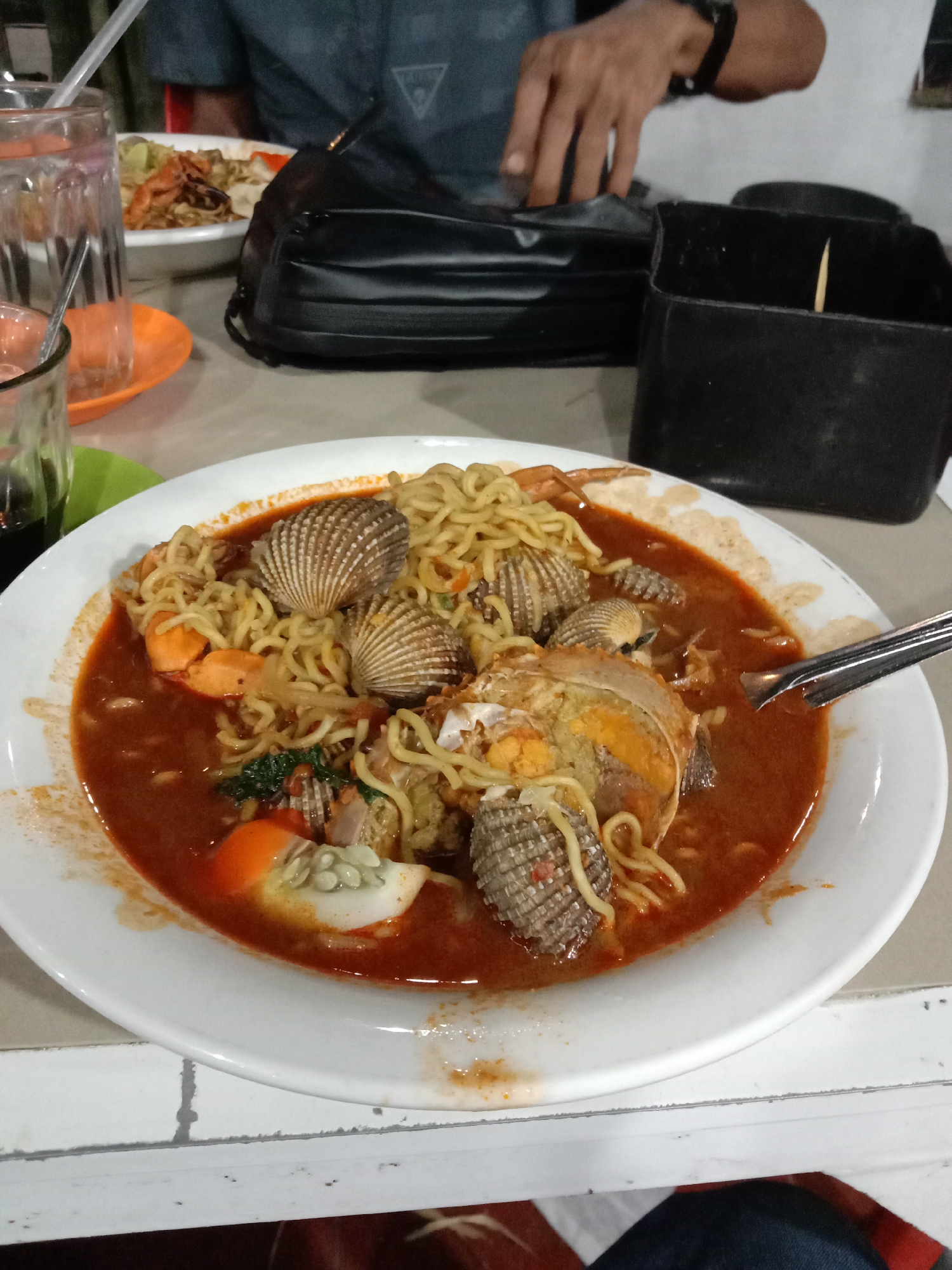 A close-up shot depicts a white plate filled with a dish consisting of noodles, seafood, and a reddish-brown broth, likely a local Indonesian cuisine. The seafood components include several cockles and a crab. The dish is garnished with green leafy vegetables and sliced eggs. A spoon rests on the plate, seemingly ready for consumption. The plate sits on a white table with a brick-like pattern.
In the background, slightly out of focus, are additional dining elements. There are two glasses, one containing a beverage and the other clear. An orange plate with food on it and a black bag. A hand is visible with a watch, partially obscured by the bag. There is also a black container with a toothpick.
The location is likely a restaurant in Lhokseumawe, Indonesia, based on the likely type of cuisine.