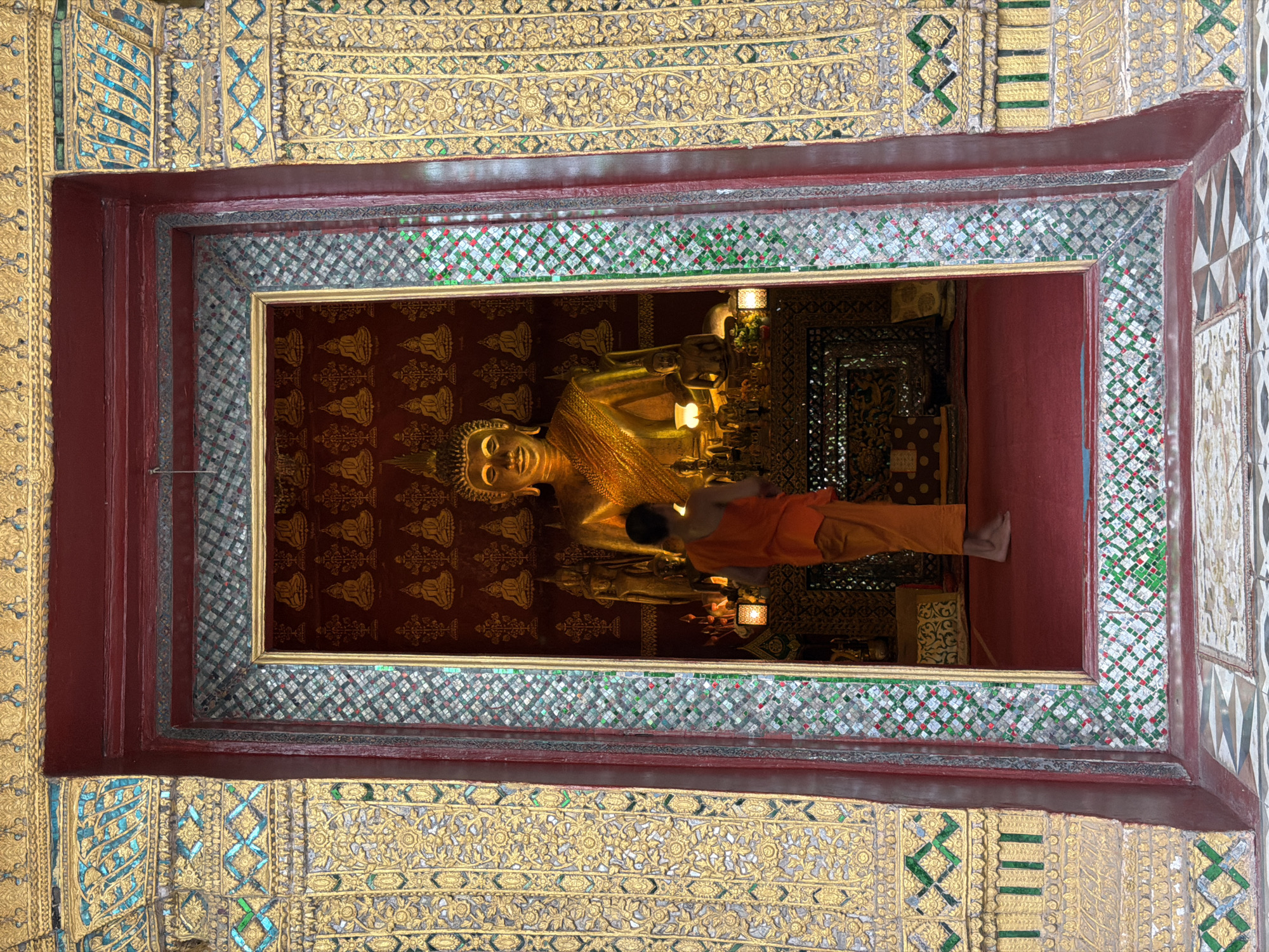 The image shows a monk standing barefoot in front of a large, golden Buddha statue inside a temple. The monk is wearing an orange robe and has his back to the camera. The Buddha statue is seated and is adorned with gold fabric. Behind the statue, there is a backdrop with a pattern of smaller Buddha images. The temple interior is richly decorated with gold and colorful mosaic tiles. The entrance to the temple is framed by ornate, gilded carvings and mosaic patterns, suggesting a significant religious or cultural site in Choumkhong, Laos. The floor inside the temple appears to be covered in a red carpet, while the floor outside the entrance has a patterned tile design.