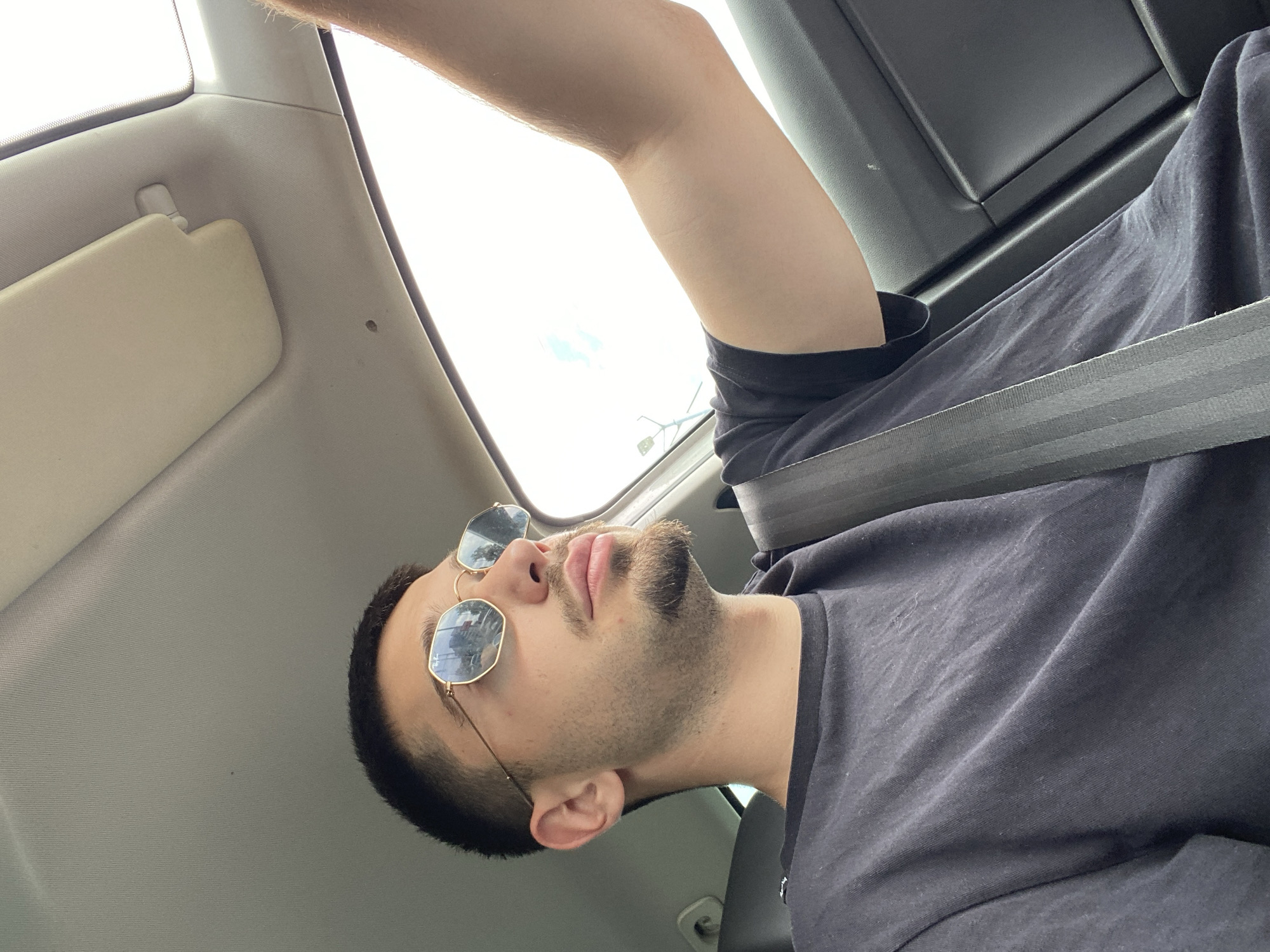 A person is sitting in a car. They have on sunglasses and are wearing a seatbelt. The car interior is visible, including the sun visor and the sunroof. The location is Plovdiv, Bulgaria, as evidenced by the reflections in the sunglasses, which show buildings and street signs with Cyrillic script. The person is angled in the seat, looking upward. The person is wearing a black t-shirt. The photograph appears to have been taken from a low angle.