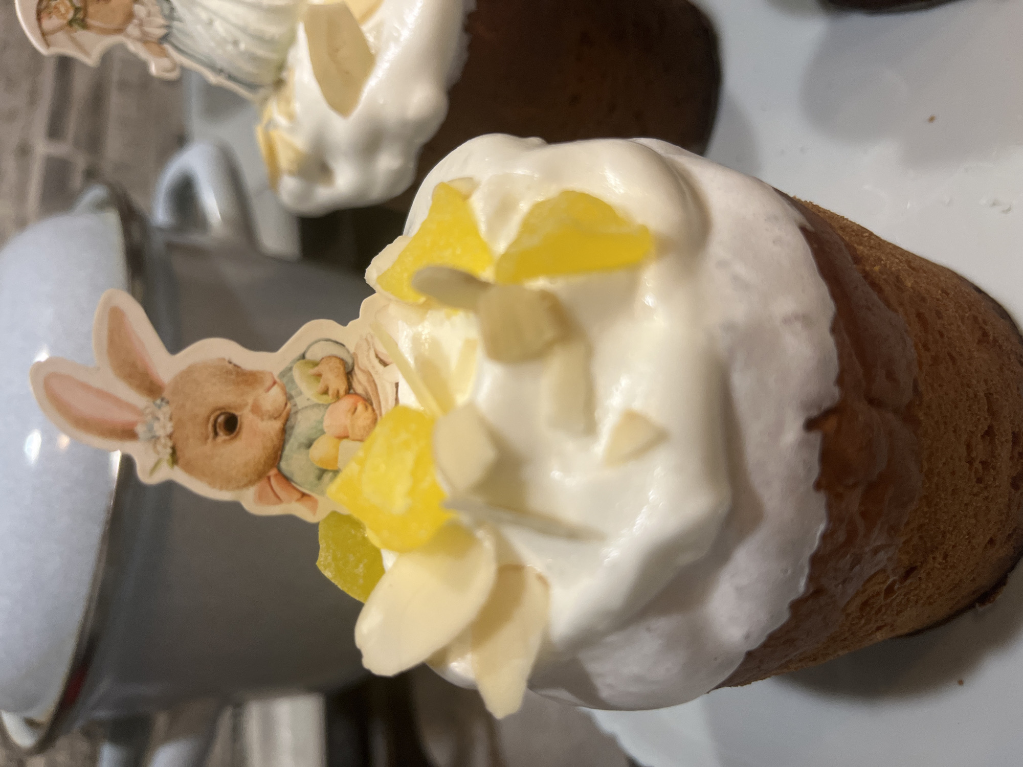 A close-up, slightly angled view shows Easter kulich bread, a traditional Russian and Ukrainian Easter bread, decorated with white frosting, diced yellow fruit (likely candied), and slivered almonds. One kulich has a bunny-shaped paper decoration inserted into the frosting. Another kulich is partially visible in the background. A metal mug or container is visible to the left. The kulich breads are presented on a white surface. The background is out of focus but appears to be an indoor setting, possibly a kitchen or dining area. There is no explicit information that places the bread in the city of Chaikovsky, Russia.