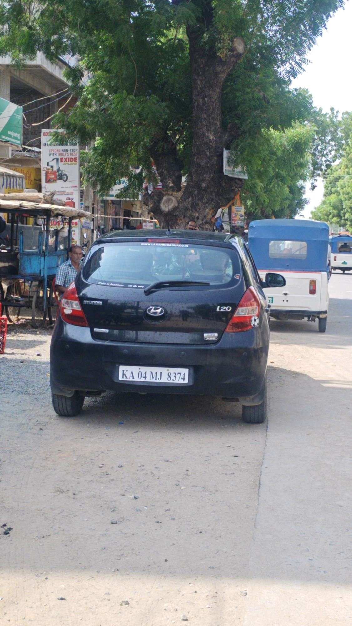 In Virat Nagar, India, a black Hyundai i20 is parked on a dusty road next to a concrete sidewalk. The car, facing away from the camera, has a license plate reading "KA 04 MJ 8374." To the left of the car, a man in a checkered shirt stands behind a blue cart with a yellow awning, possibly a food or goods vendor. Behind the car, a blue auto-rickshaw is visible, followed by another white auto-rickshaw. A large tree with dense green foliage dominates the background, with some buildings and signage visible to the left. The overall environment suggests a roadside scene in a moderately busy urban area.