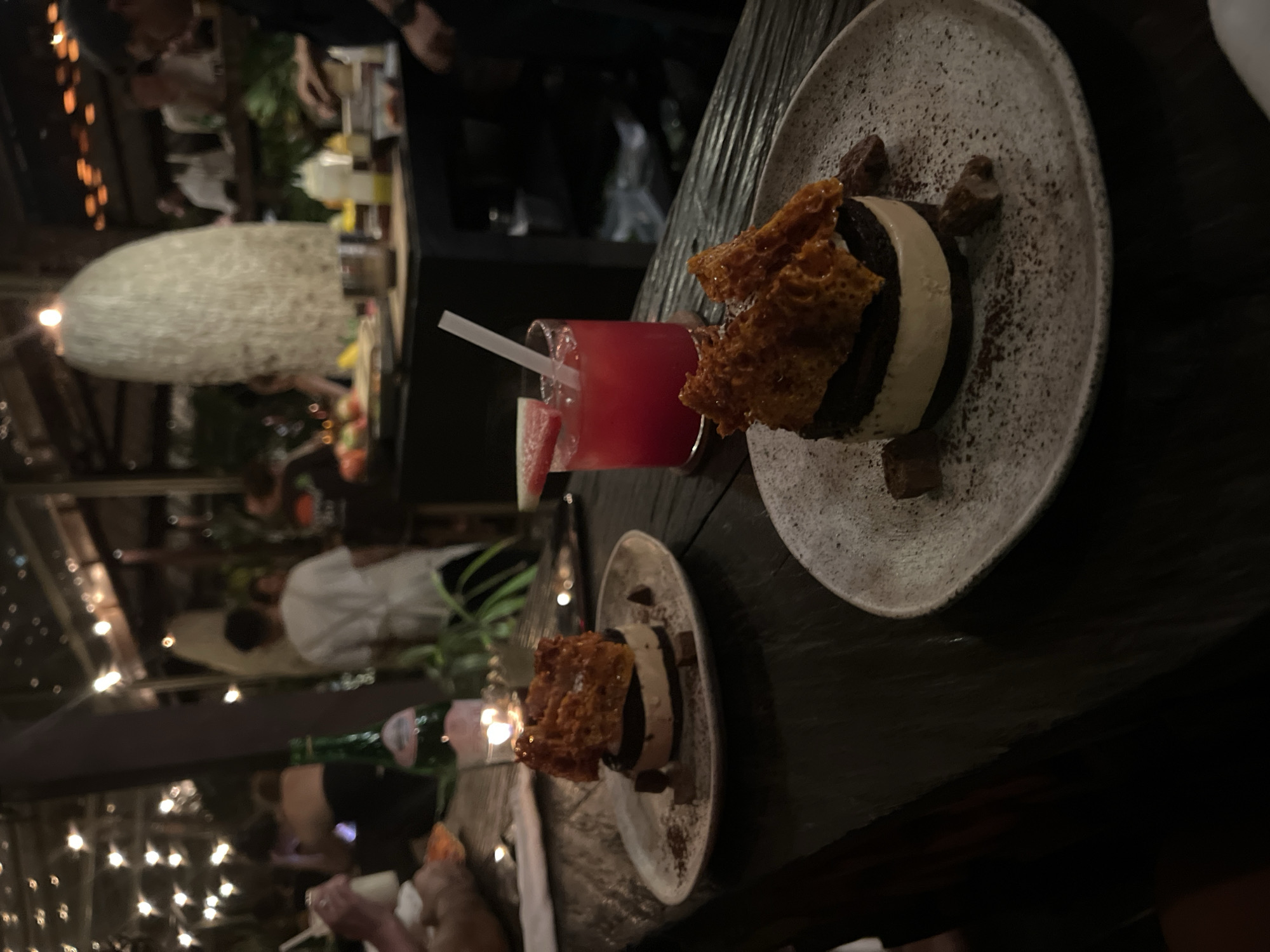 The image shows a nighttime scene in Mengwi, Indonesia, likely at a restaurant or bar. There are at least two plates of desserts visible on a dark wooden table. The desserts appear to be stacked discs of chocolate and cream, topped with a crispy, golden-brown honeycomb-like structure. Cocoa powder is sprinkled around the desserts on the plates. Next to the desserts are glasses of a pink-red beverage with watermelon slices and white straws. Behind the table, there are people blurred in the background, some wearing white shirts. String lights and a large, white, woven light fixture add to the ambiance. A bottle of water is also present.