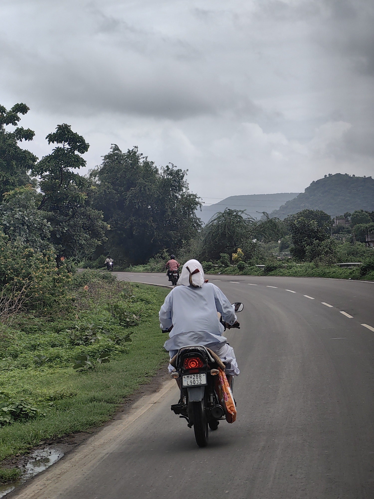The image shows a person riding a motorcycle on a road in Rajur, India. The rider is wearing a light blue garment and a white head covering. The motorcycle has a license plate that reads "MH28B L9032". The road is paved and has white dashed lines indicating lanes. On the left side of the road, there is a grassy area with lush green vegetation and trees. In the background, there are rolling hills covered with trees, and the sky is overcast. Another motorcycle with a rider in a red shirt is visible further up the road. The scene appears to be a rural or semi-rural setting.
