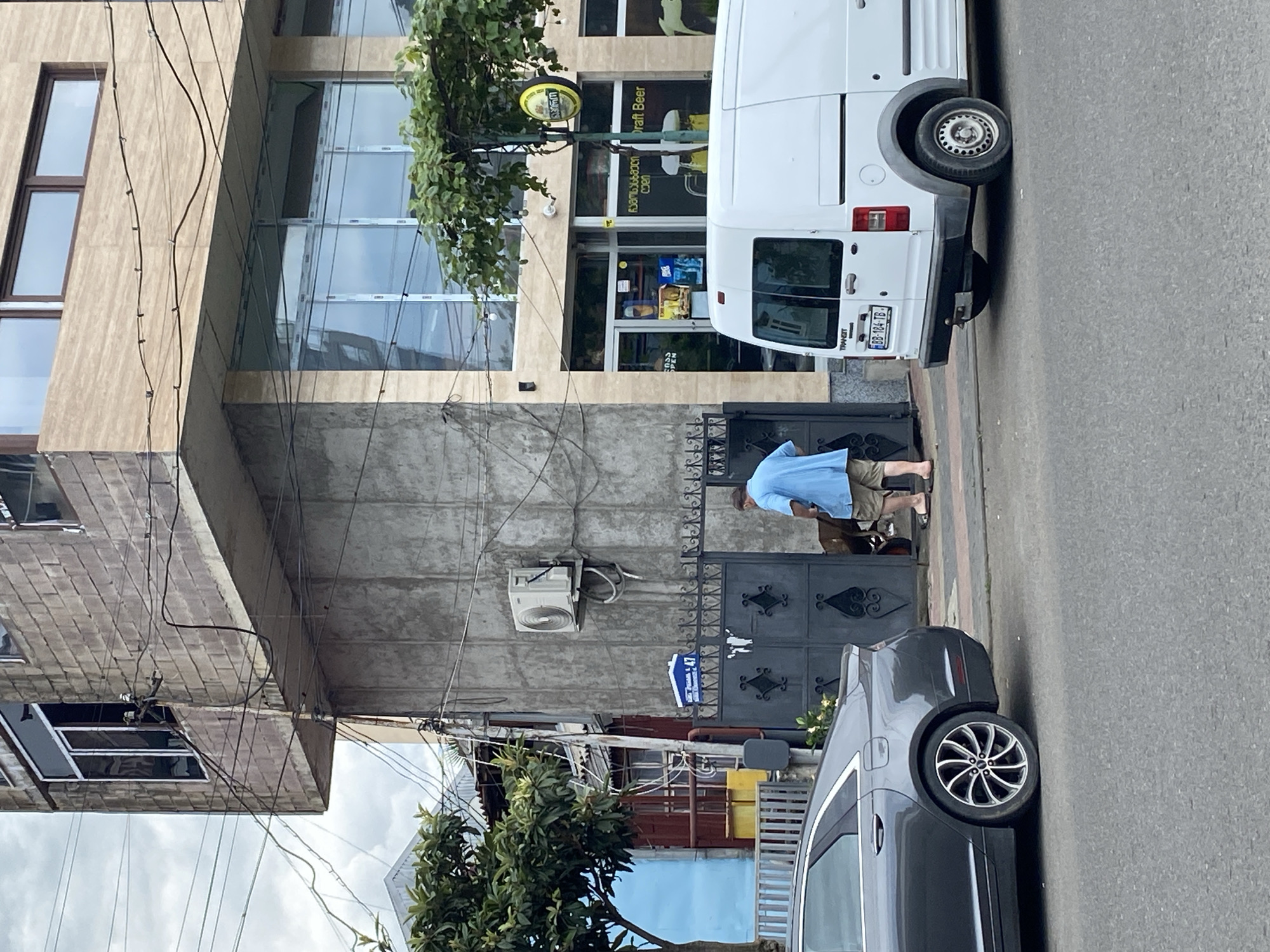 The image captures a street scene in Batumi, Georgia. In the foreground, a dark gray sedan is parked on the left, facing away from the camera. To its right, a man wearing a light blue shirt and tan shorts is standing near a dark metal gate. He appears to be interacting with something or someone within the gated area, possibly a dog that is partially visible. Behind the man and the gate, a white van is parked on the right side of the street, facing away from the camera. The street is paved with asphalt.
The background features a multi-story building with a modern facade, characterized by large windows and a mix of concrete and beige tiled surfaces. An air conditioning unit is mounted on a concrete section of the building. Above the gate, a blue street sign with white lettering is visible. Further back, there are glimpses of other buildings and trees, suggesting an urban environment with residential and commercial structures. The sky appears overcast. The overall impression is a candid snapshot of everyday life on a Batumi street.