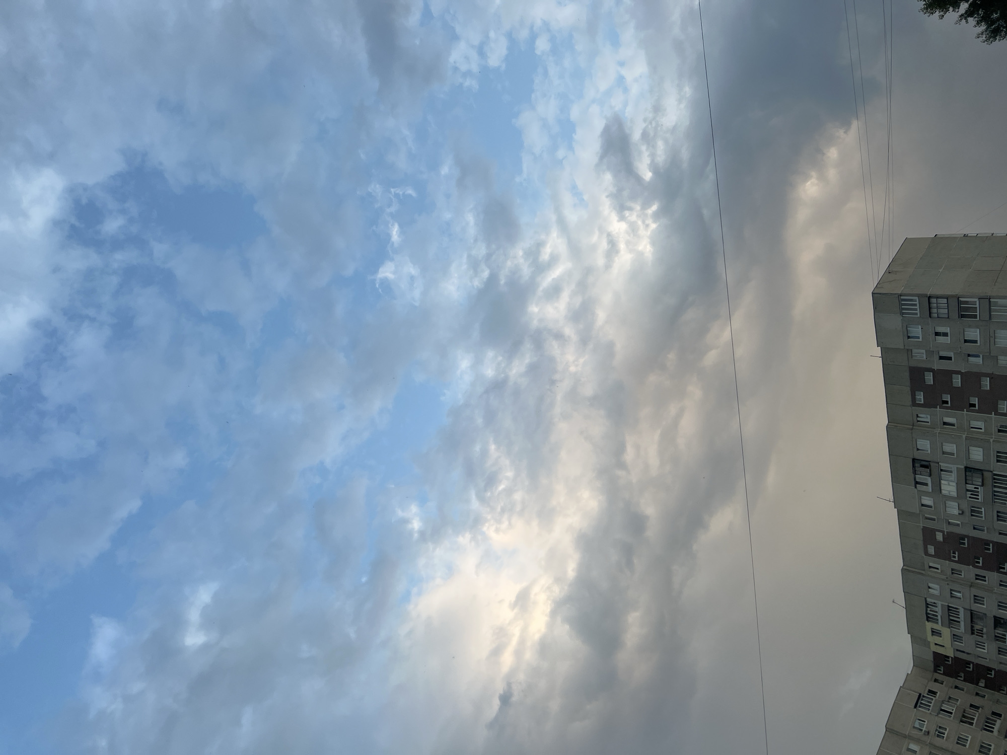 An eye-level, vertical, medium shot captures the sky above a multi-story residential building in Ulyanovsk, Russia. The sky is a canvas of soft blues and grays, with clouds interspersed throughout. Some clouds are light and wispy, while others are denser and darker, hinting at impending weather changes. A faint line, possibly a wire, stretches horizontally across the mid-ground of the image. The lower portion of the frame is dominated by the side of a plain, gray apartment building. The building has numerous windows, some open and others closed, and balconies. A small portion of a dark green tree is visible on the far right edge of the frame. The scene is devoid of any people or overt actions.