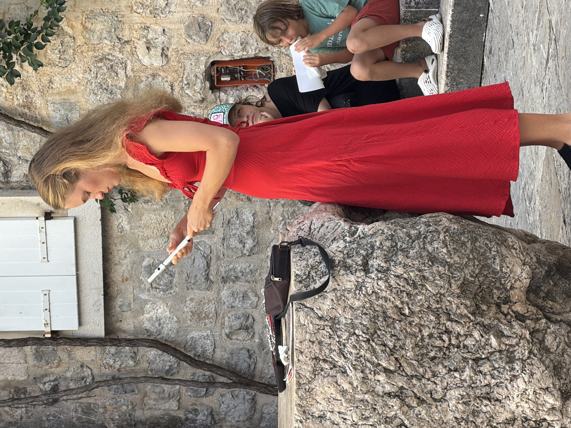 A young woman with long blonde hair, wearing a red dress, stands outdoors in a city, holding a flute. She is on a stone surface in front of a stone wall with white shutters. To her right, a young boy in a green shirt and red shorts sits on stone steps, eating from a white container. Behind the woman, another person wearing a black shirt and a green and white baseball cap looks on. An electrical box is visible on the stone wall behind them. The scene appears to be in Budva, Montenegro, possibly near the Old Town given the stone architecture.