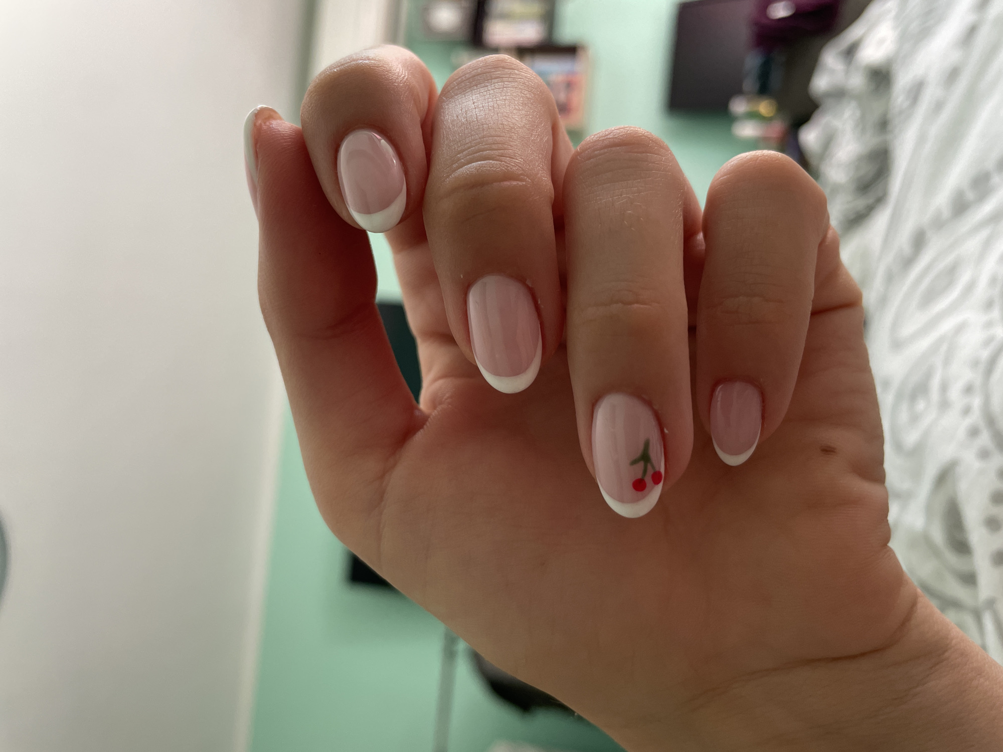 A close-up shot focuses on a person's left hand, displaying fingernails adorned with a classic French manicure. The nails are oval-shaped with a light pink base and crisp white tips. Notably, one nail features a small, playful design of two red cherries with a green stem. The hand is positioned in a way that highlights the nail art. The background is blurred, showing a mint green wall and a bed with white bedding, suggesting an indoor residential setting. The lighting appears to be natural daylight, casting soft shadows. The image's context within a city is unknown.